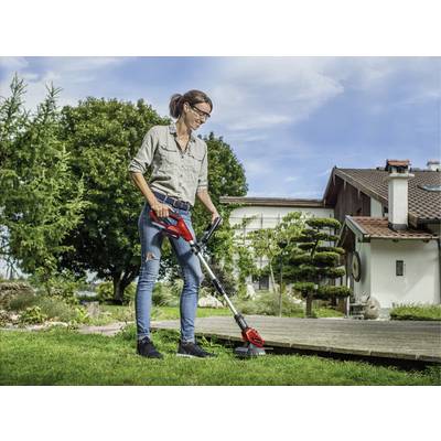 Žena používa krovinorez v záhrade vedľa domu. Má na sebe džínsy a košeľu. V pozadí sú stromy a modrá obloha.