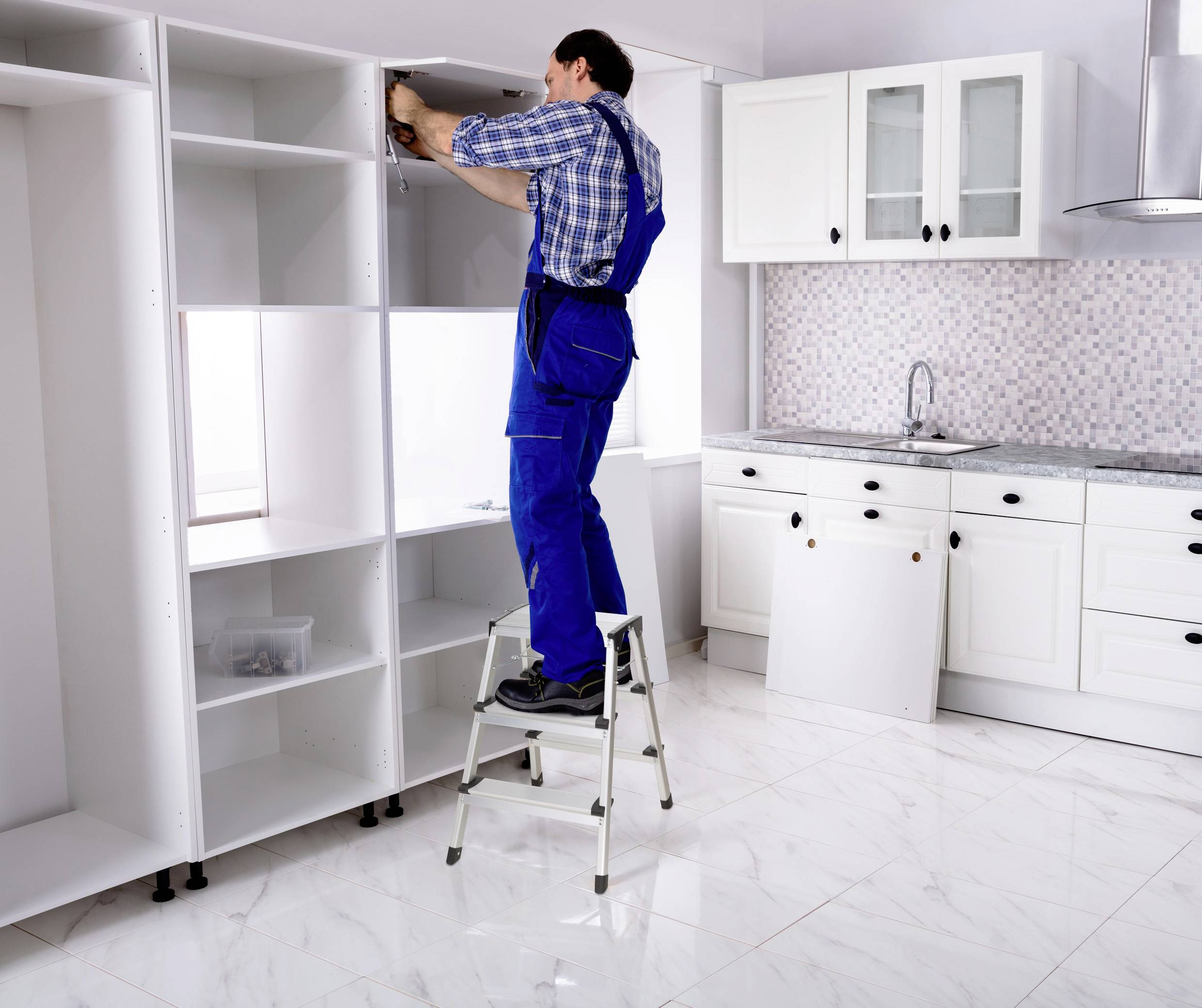 Muž v pracovnom oblečení stojí na rebríku a montuje kuchynský nábytok v bielej kuchyni.