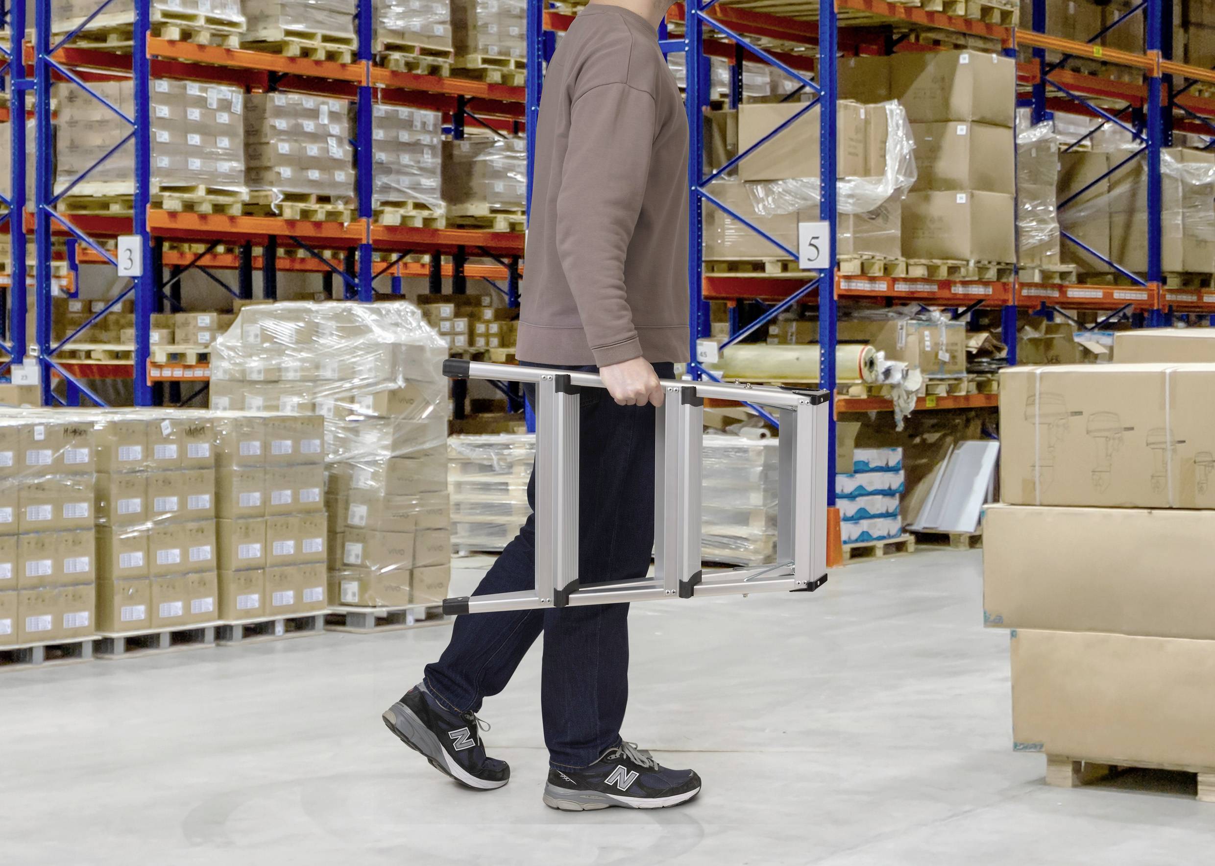 Osoba nesie rebrík cez sklad s paletami a regálmi plnými krabíc a balíčkov v pozadí.