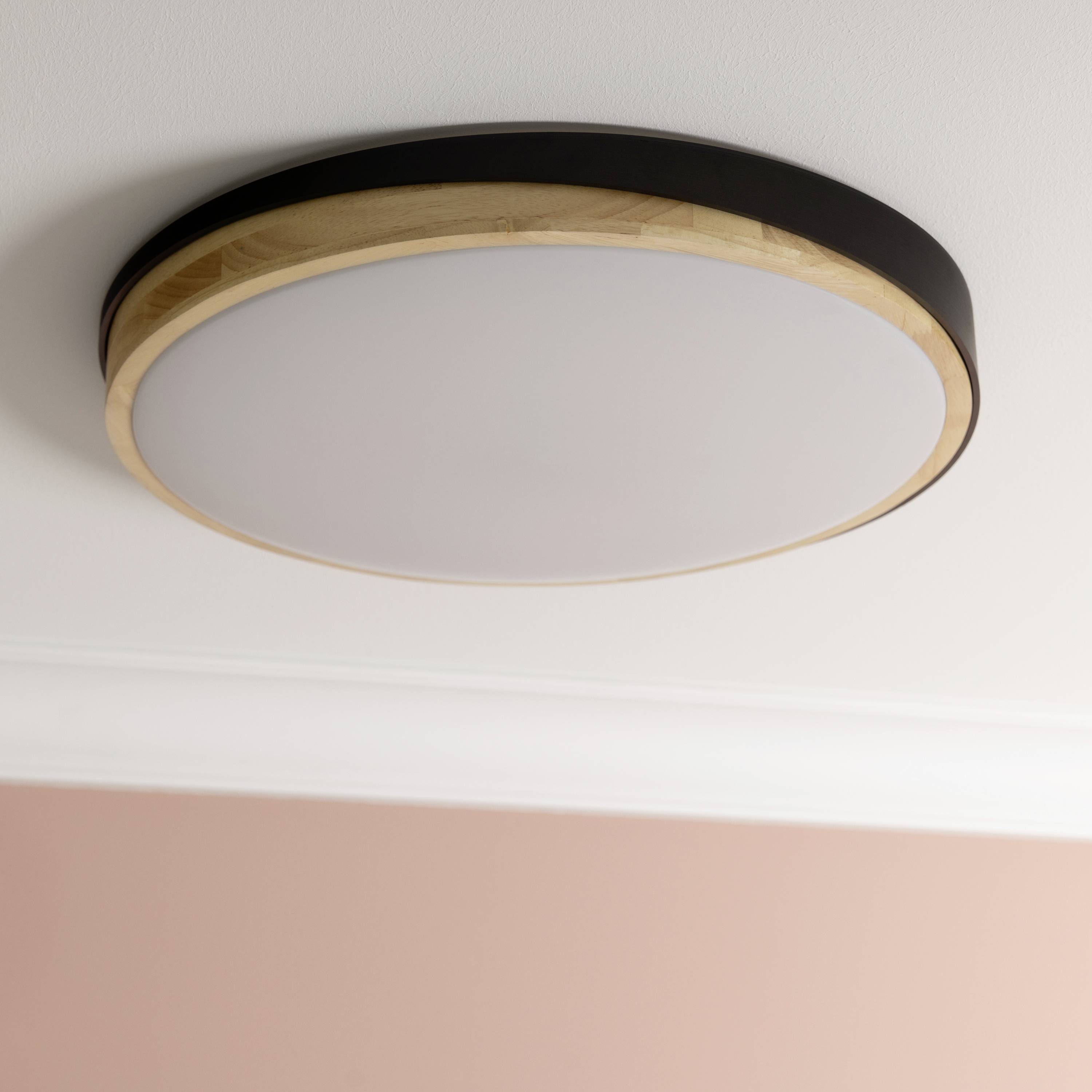 Okrúhle stropné svietidlo s prvkami z dreva a kovu, namontované na bielom strope. Pod ním sa nachádza ružová stena.