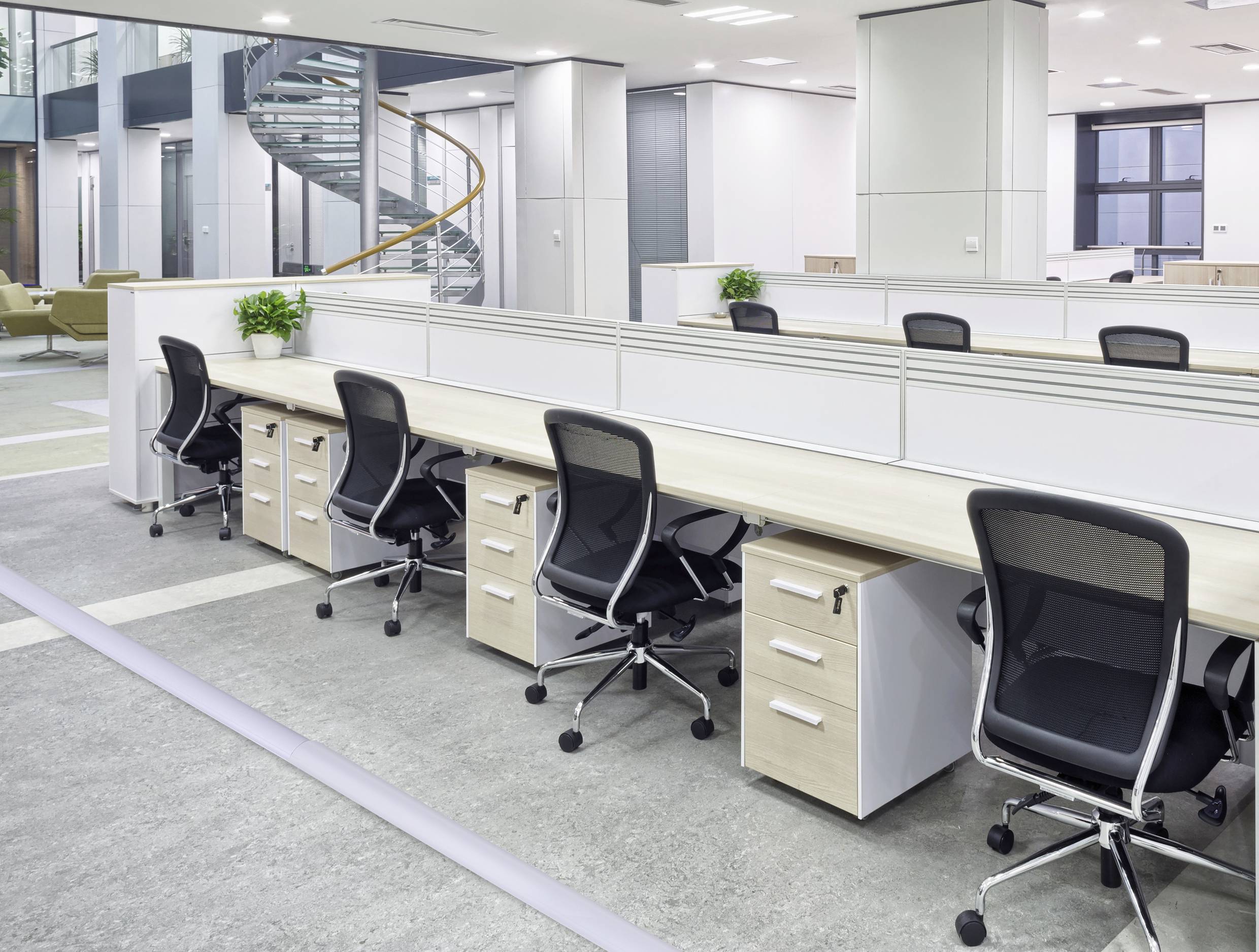 Veľkopriestorová kancelária s niekoľkými prázdnymi písacími stolmi a kancelárskymi stoličkami, jasne osvetlená, s modernými nábytkom a točitým schodiskom v pozadí.