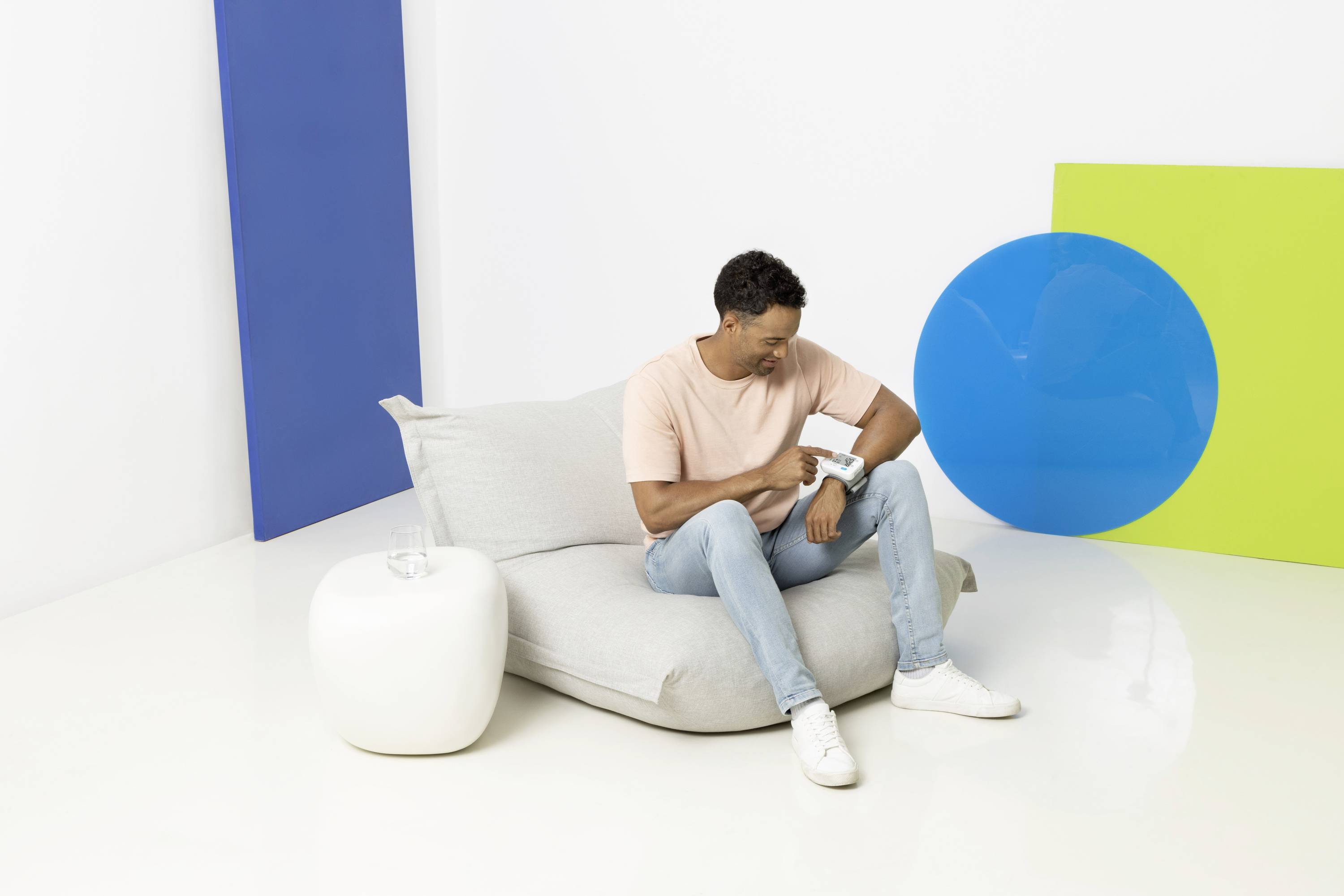 Muž sedí na sivom sedacom vaku v modernej, farebnej miestnosti a pozerá na zariadenie na svojom zápästí.