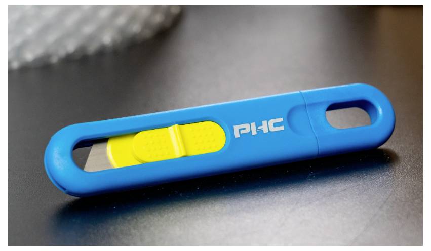 Modrý, obdĺžnikový posúvač so žltým prepínačom, ktorý leží na sivom povrchu. Na posúvači je napísané 'PHC'.