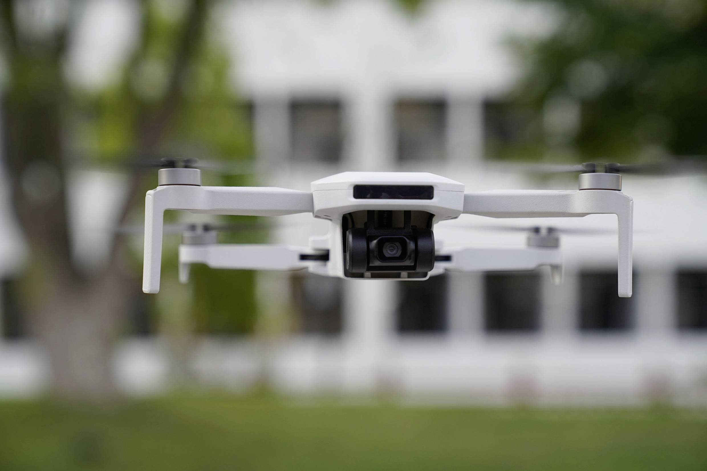 Biela dron vznáša vo vzduchu pred rozmazaným pozadím s postavenými stromami a viacposchodovou budovou.