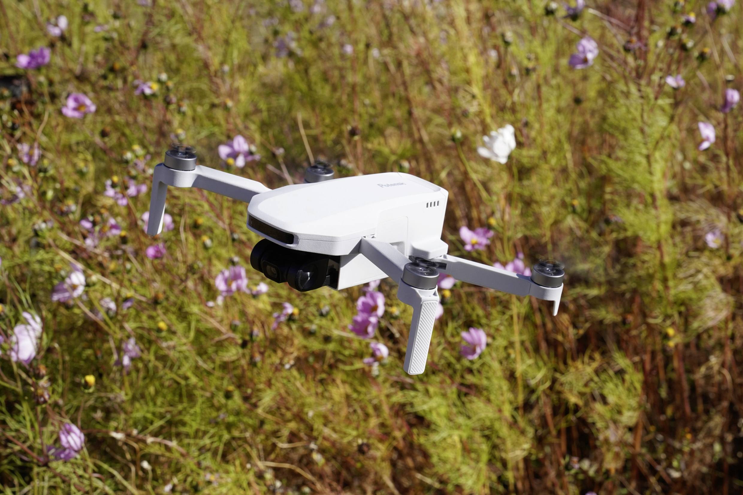 Biela dron letí nad poľom s kvitnúcimi rastlinami v pozadí.