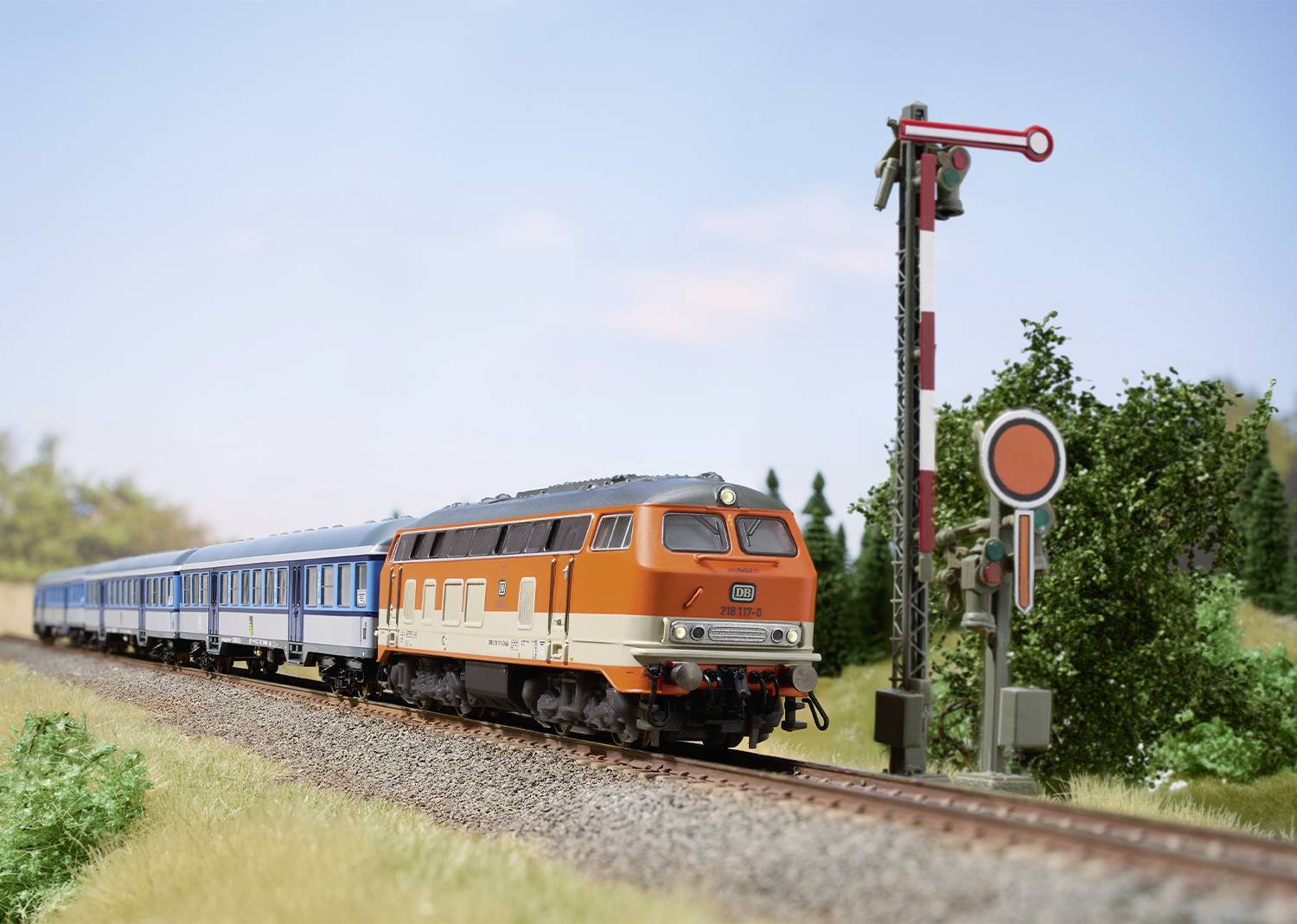 Oranžovo-modrý vlak ide po železničnej trati cez zelenú krajinu. Mechanický signál je nastavený na zastavenie.