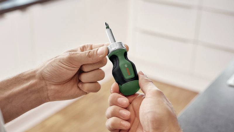 Detailný záber osoby držiacej skrutkovač so zelenou rukoväťou. V rozmazanom pozadí je vidieť kuchyňu.