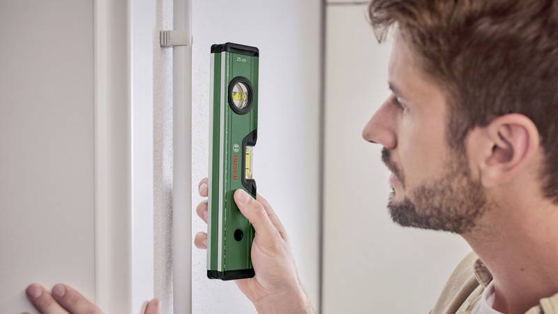 Muž vyrovnáva vodováhu vertikálne na stene, aby skontroloval rovnosť. Vodováha ukazuje horizontálnu bublinu.