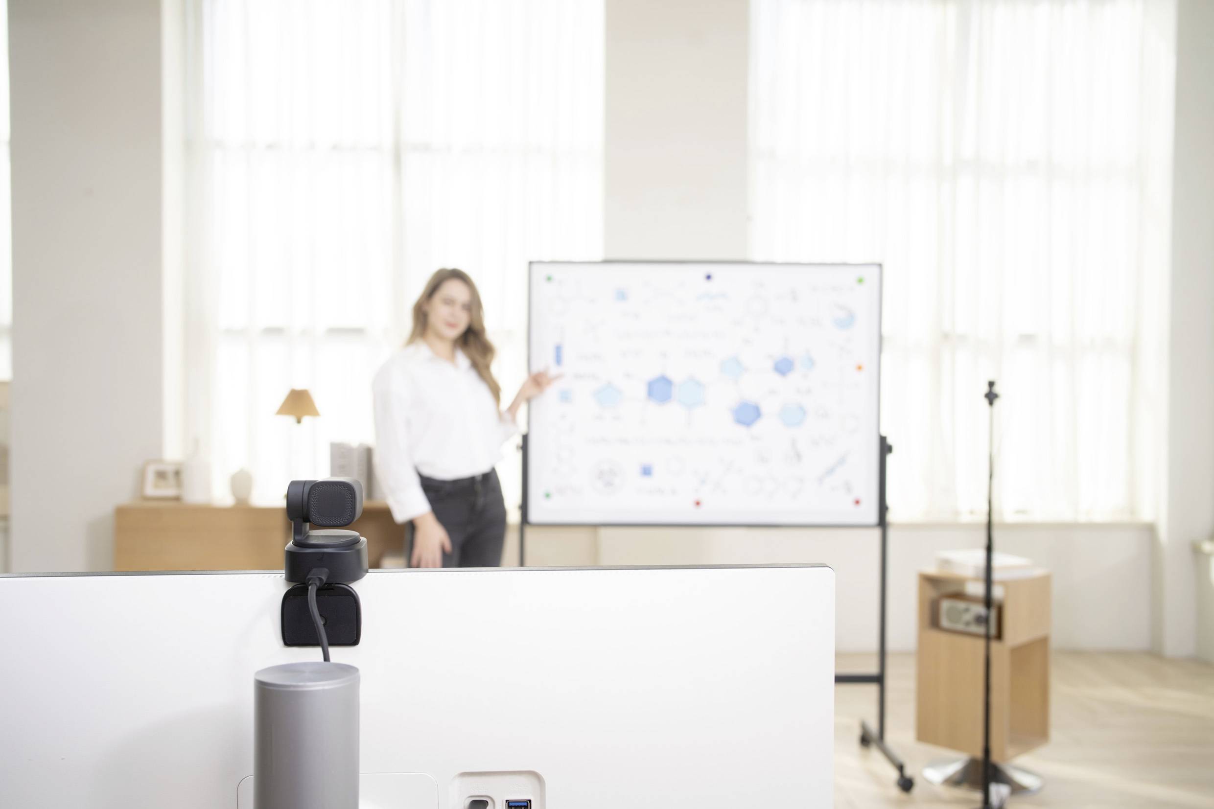 Žena ukazuje na whiteboard s diagramami a výkresmi vo svetlej miestnosti, pričom kamera je pripevnená na monitore.