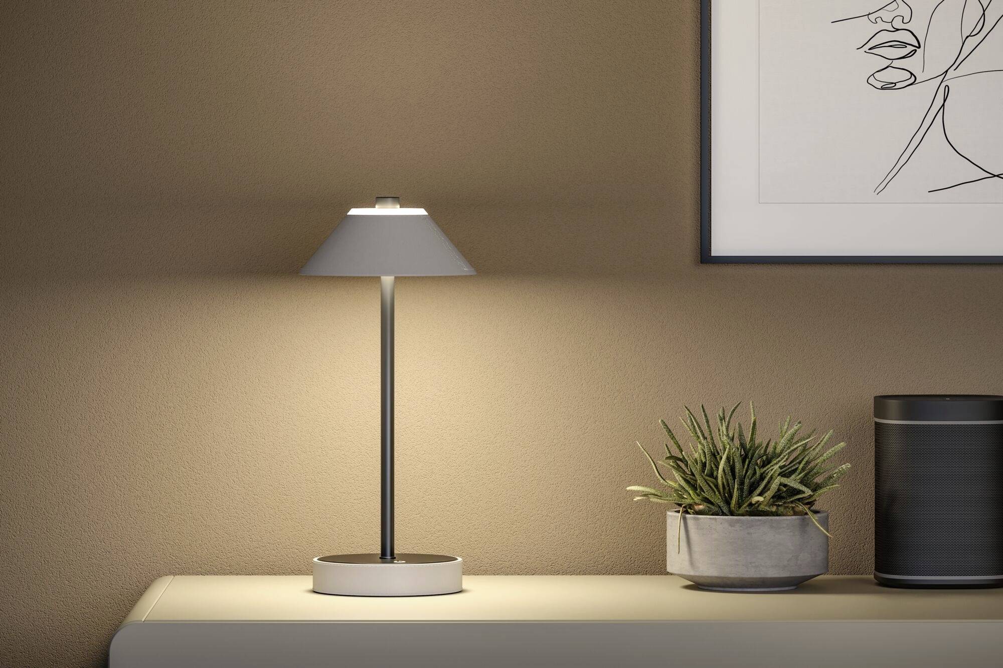 Moderná stolová lampa svieti na svetlom vedľajšom stolíku vedľa rastliny. Na stene visí minimalistické umelecké dielo.