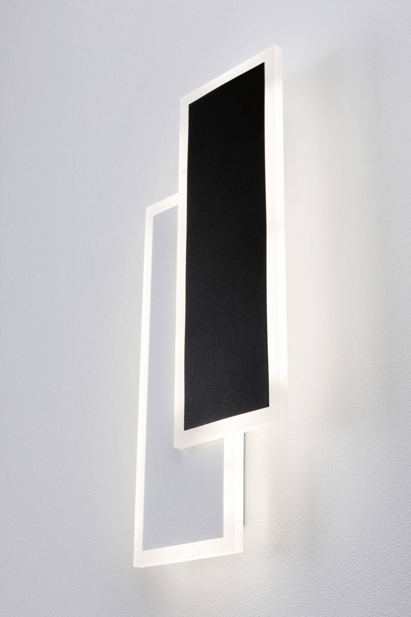 Moderná nástenná lampa s dvoma obdĺžnikovými prvkami uloženými nad sebou, jeden v čiernej a druhý v bielej farbe, svieti na stene.