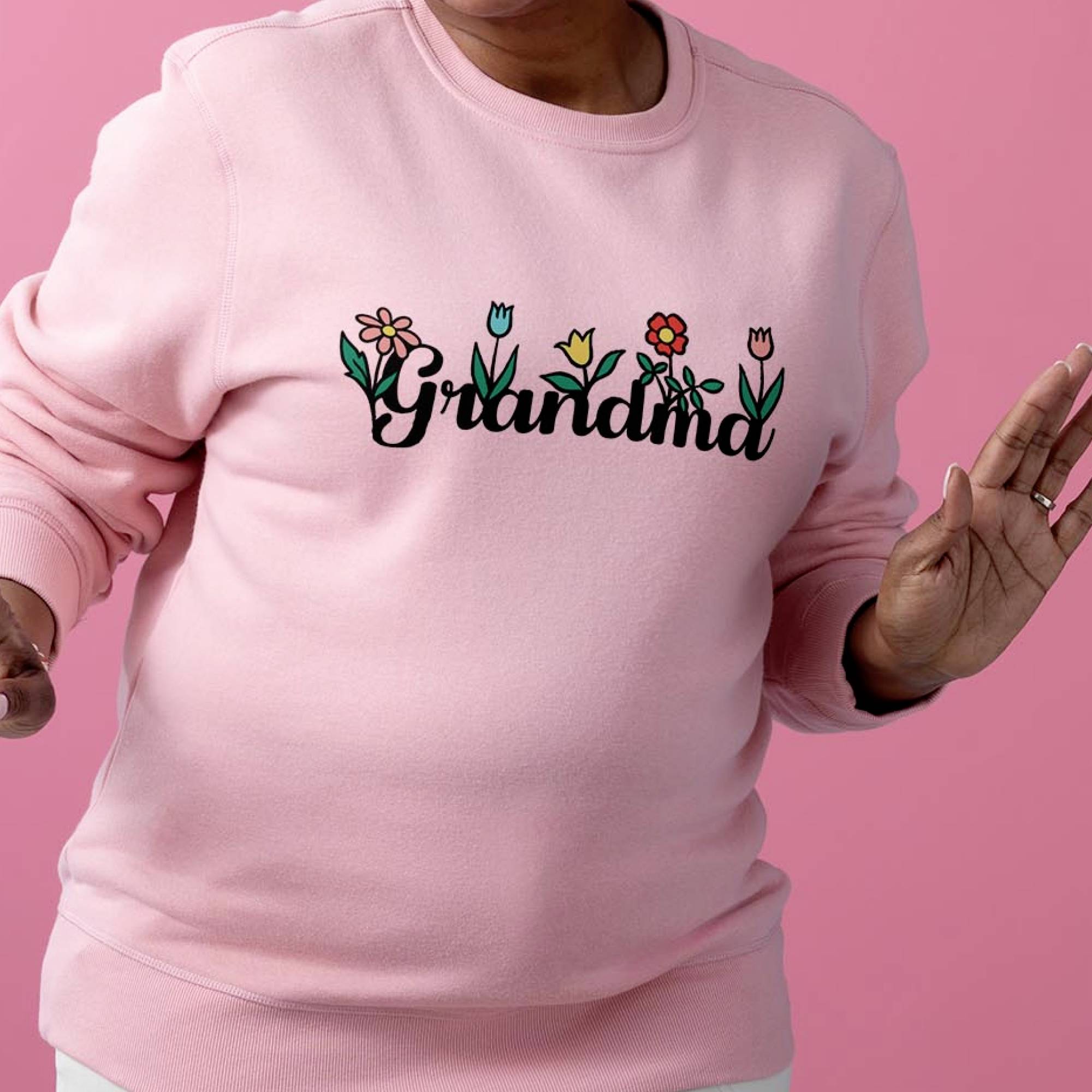 Osoba má na sebe ružový pulóver s nápisom 'Grandma' a kvetinovými vzormi pred ružovým pozadím.