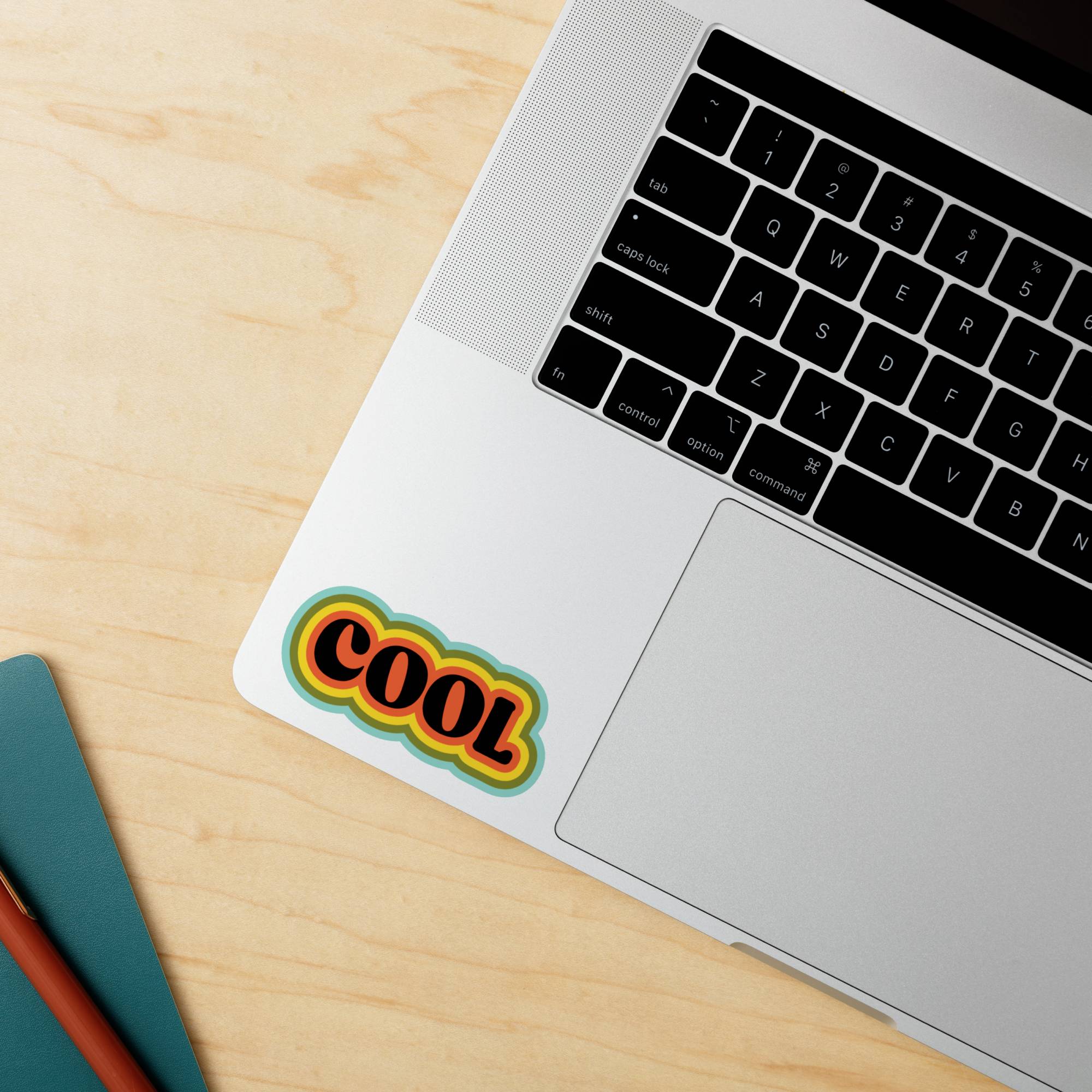 Laptop na drevenej doske so živofarebnou nálepkou s nápisom 'COOL' vedľa klávesnice.
