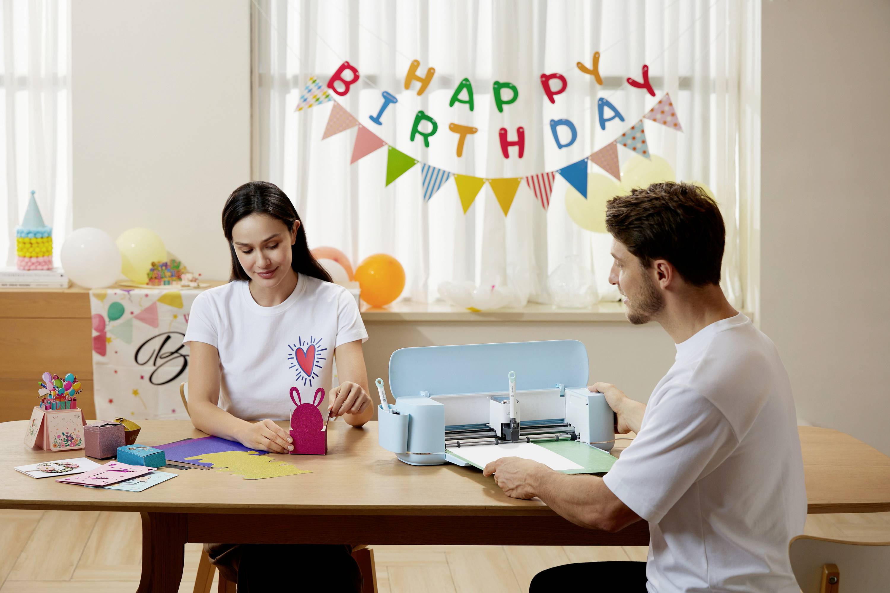 Žena a muž vyrábajú birthday dekorácie pomocou rezačky. V pozadí visí banner s nápisom "Happy Birthday".