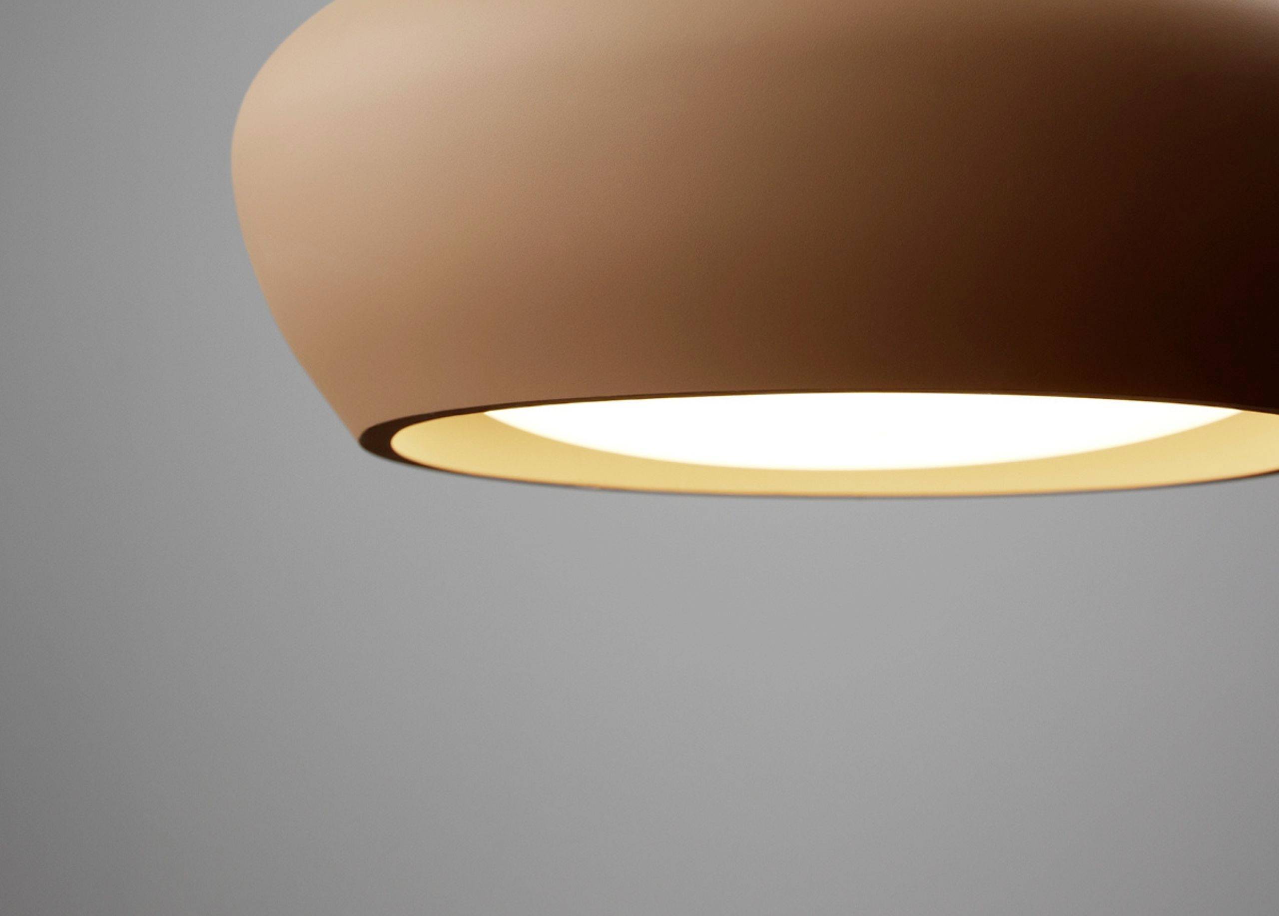 Moderná hnedá závesná lampa s teplým svetlom, ktoré svieti zhora nadol na hladkú sivú stenu v pozadí.