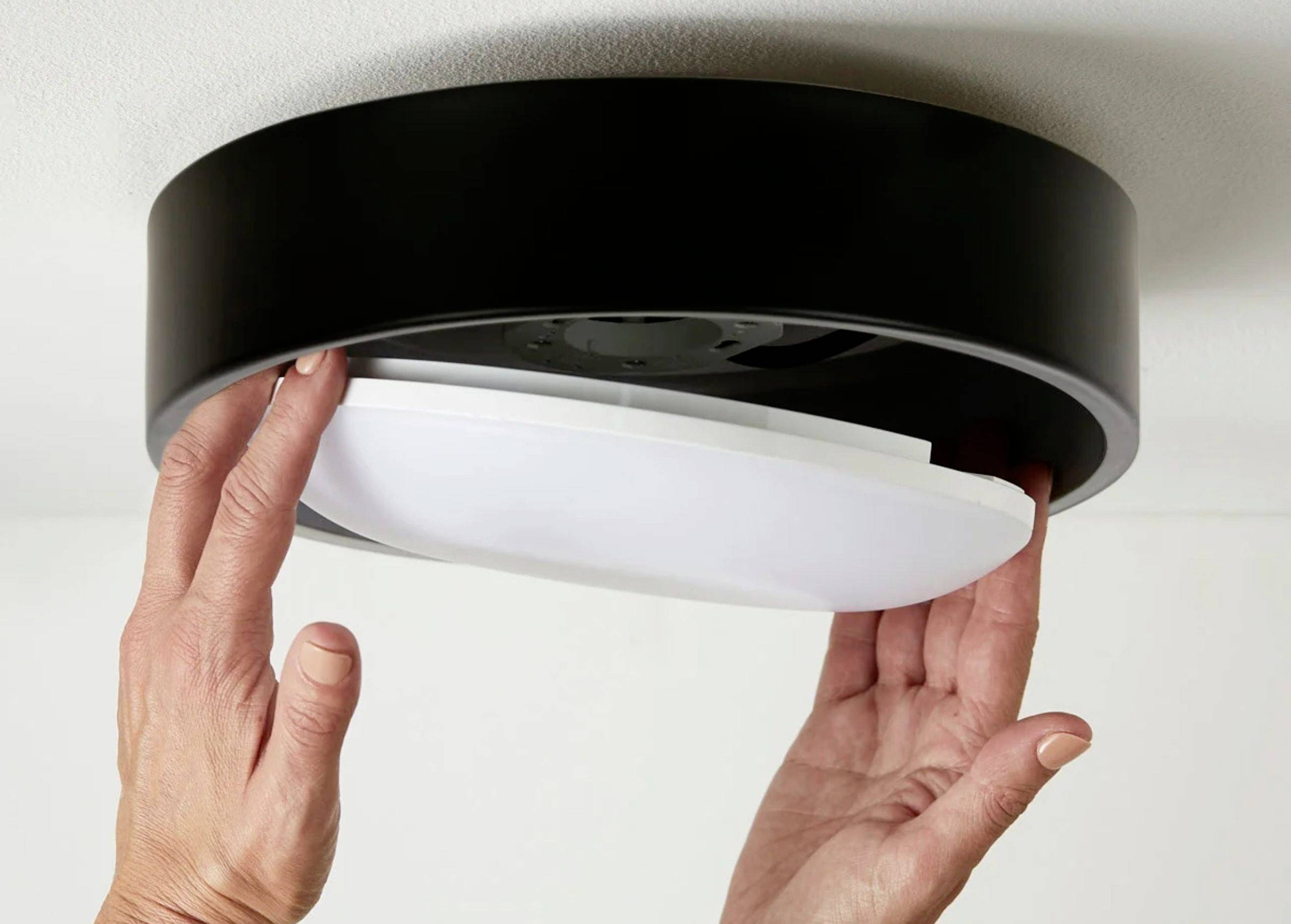 Osoba inštaluje okrúhle stropné svetlo. Ruky držia biely tienidlo, ktoré zapadá do čierneho púzdra.