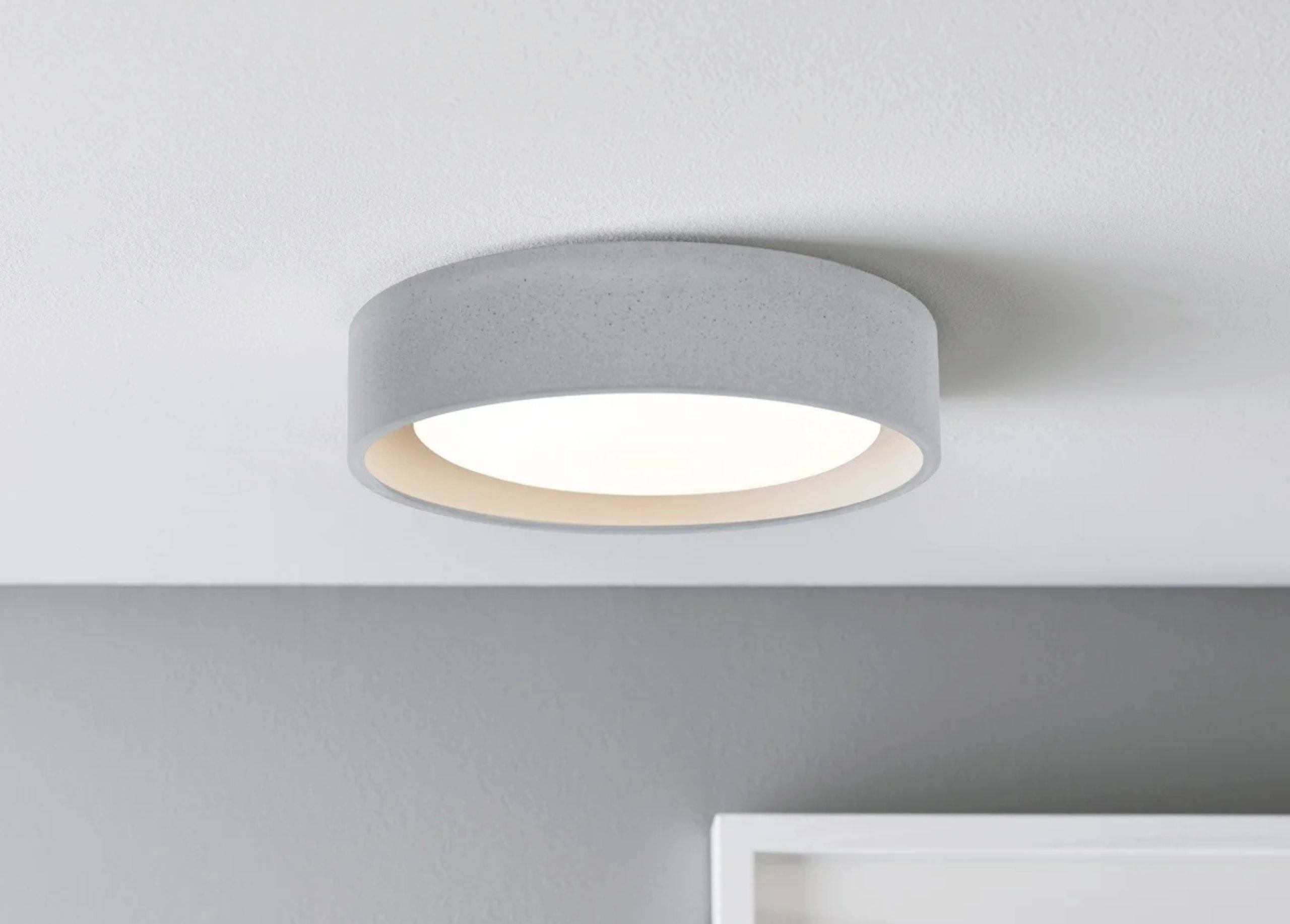 Okruhla, plochá stropná lampa so sivým rámom na svetlosivom strope, ktorá vyžaruje mäkké, rovnomerné svetlo.