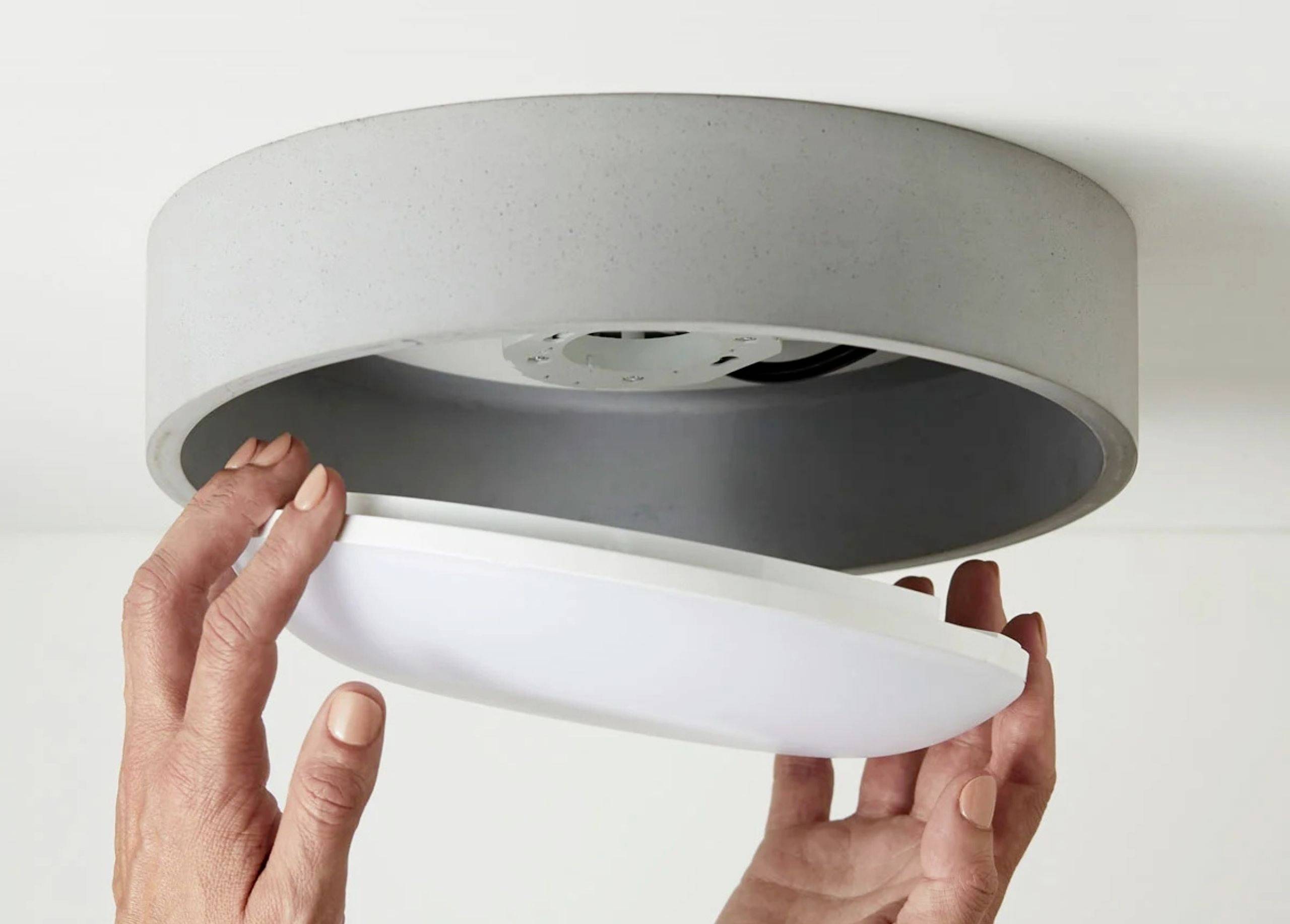 Osoba inštaluje okrúhle stropné svietidlo. Ruky pridržiavajú spodnú časť svietidla. Svietidlo má jednoduché, moderné dizajnové prevedenie.