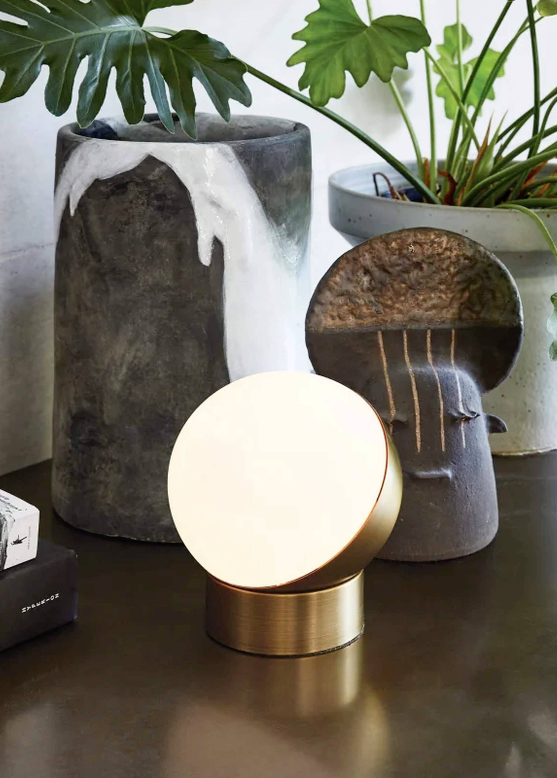 Moderná stolná lampa s okrúhlym, svietiacim tienidlom na bronzovom podstavci. Stojí pred minimalistickými, dekoratívnymi vázami a rastlinami.
