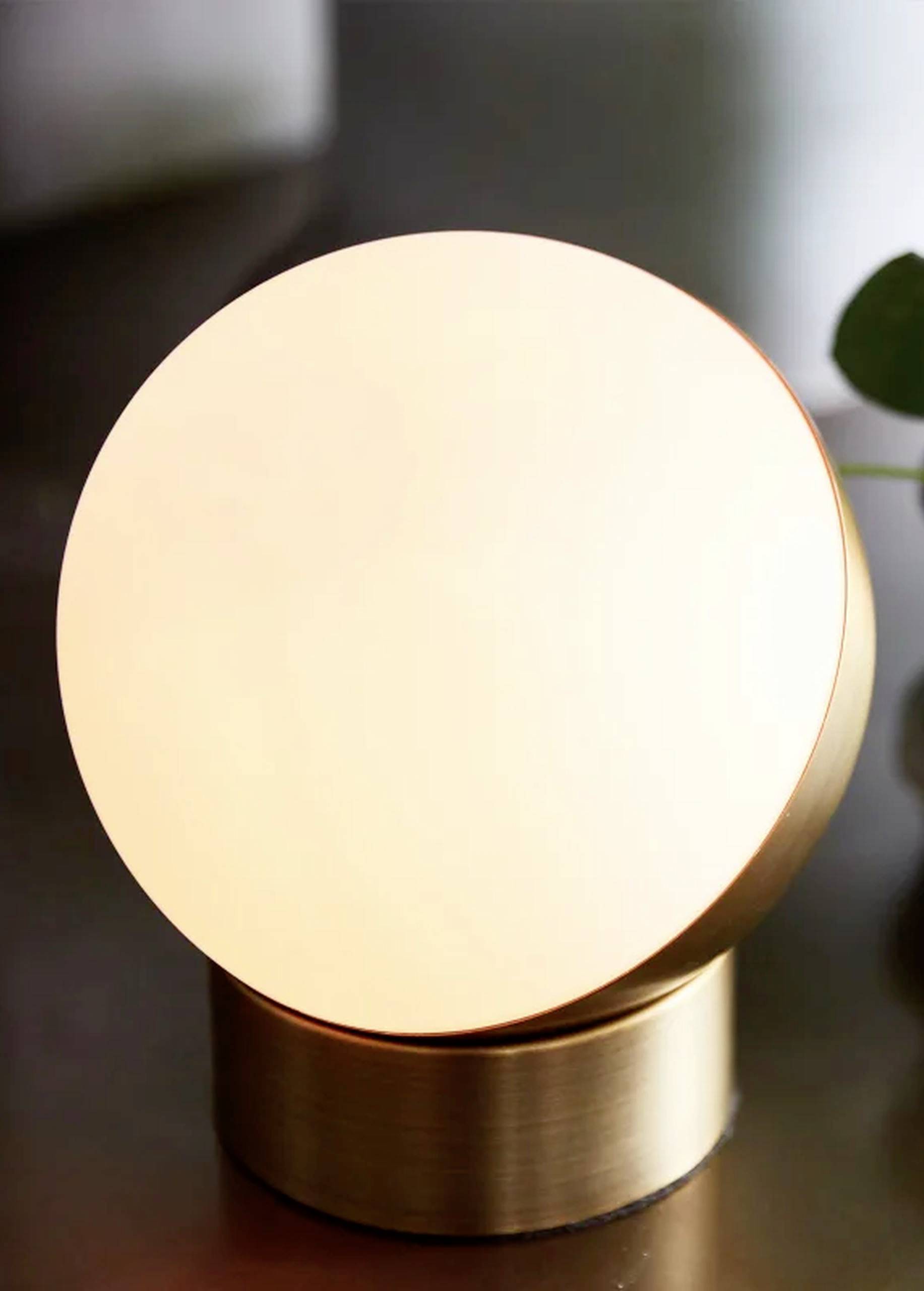 Okrúhla zlatá stolná lampa so svetlým, rozptýleným svetlom na tmavom pozadí. V pozadí je viditeľný list rastliny.