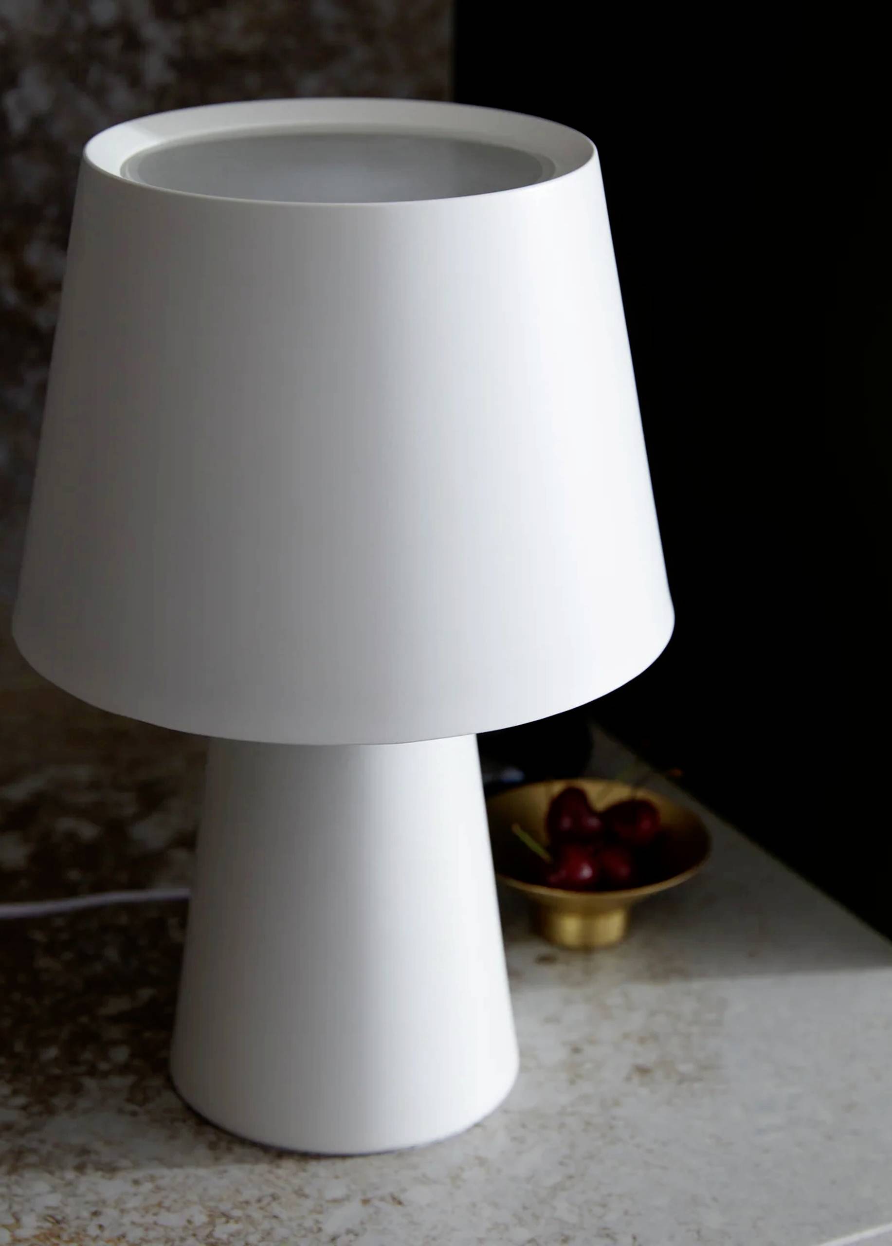 Jednoduchá biela stolová lampa na stole, vedľa nej malá miska s čerešňami.