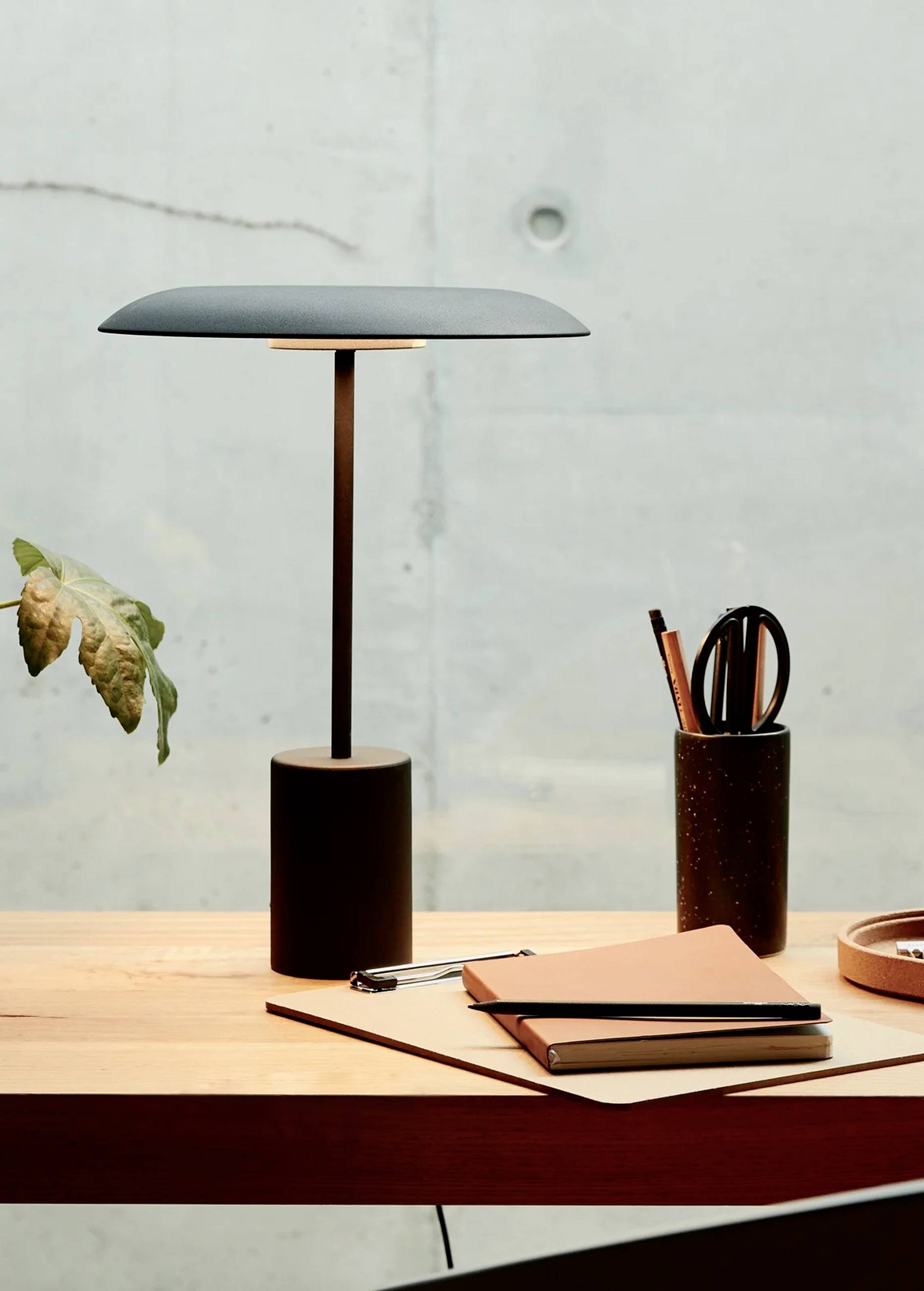 Moderná stolná lampa s plochým tienidlom osvetľuje písací stôl, na ktorom sú poznámkové zošity, perá a rastlina.