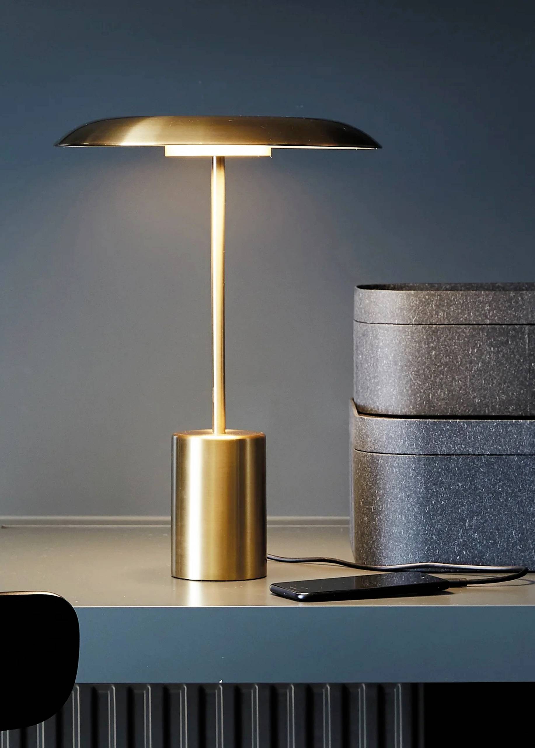 Zlatá stolná lampa svieti na písacom stole. Vedľa nej leží smartfón a stoja dva sivé reproduktory.