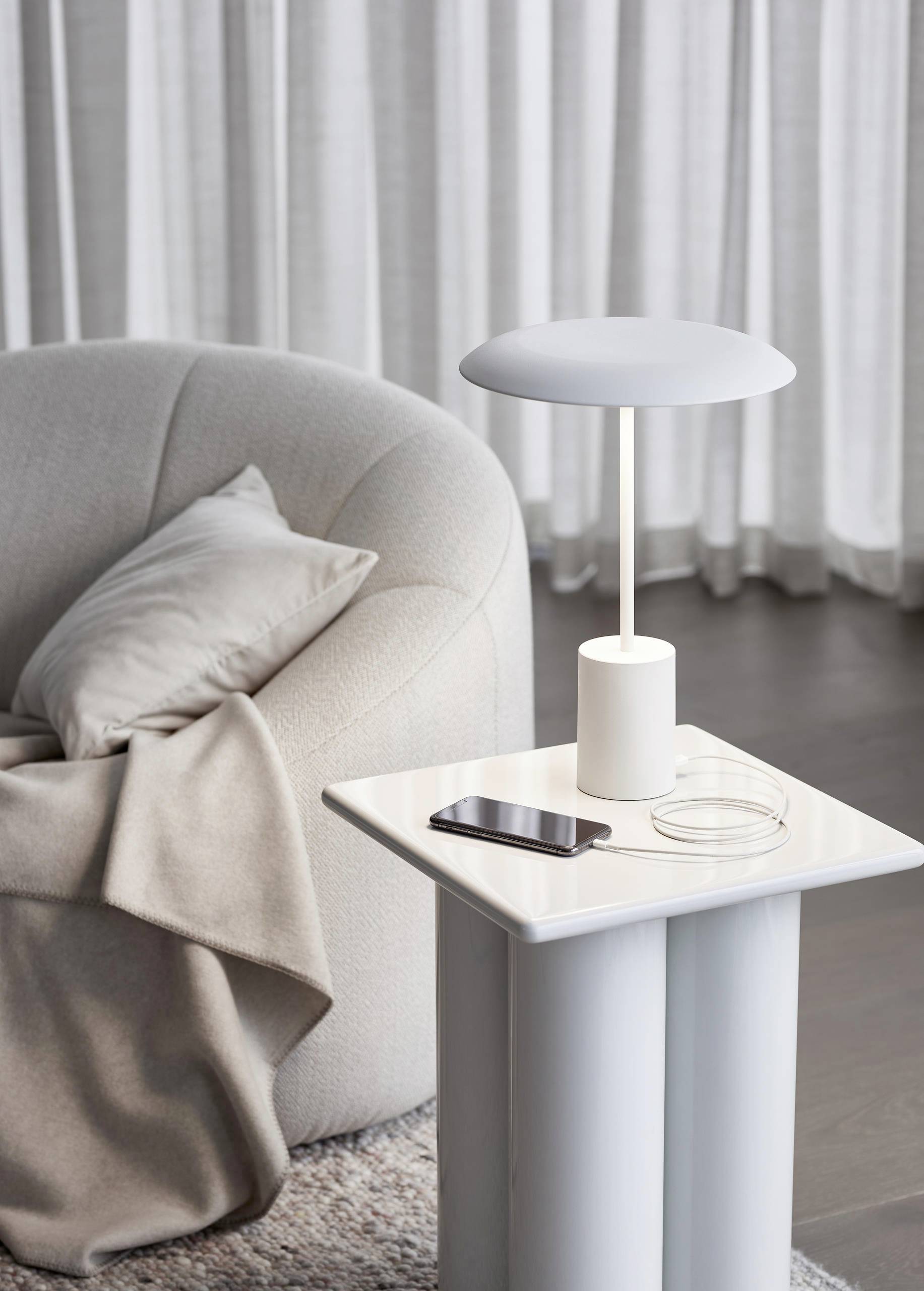 Biela lampa a smartfón na modernom príručnom stolíku vedľa pohodlného kresla s vankúšom a dekou, pred záclonami siahajúcimi až po podlahu.