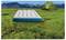 Biely nafukovací matrac na zelenej tráve so zeleným stanom v pozadí, zasadený do horskej krajiny pod jasne modrou oblohou.