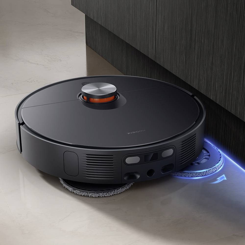 Čierny robotický vysávač čistí podlahu vedľa tmavej drevenej skrinky. Pod zariadením svieti modrá LED dióda.