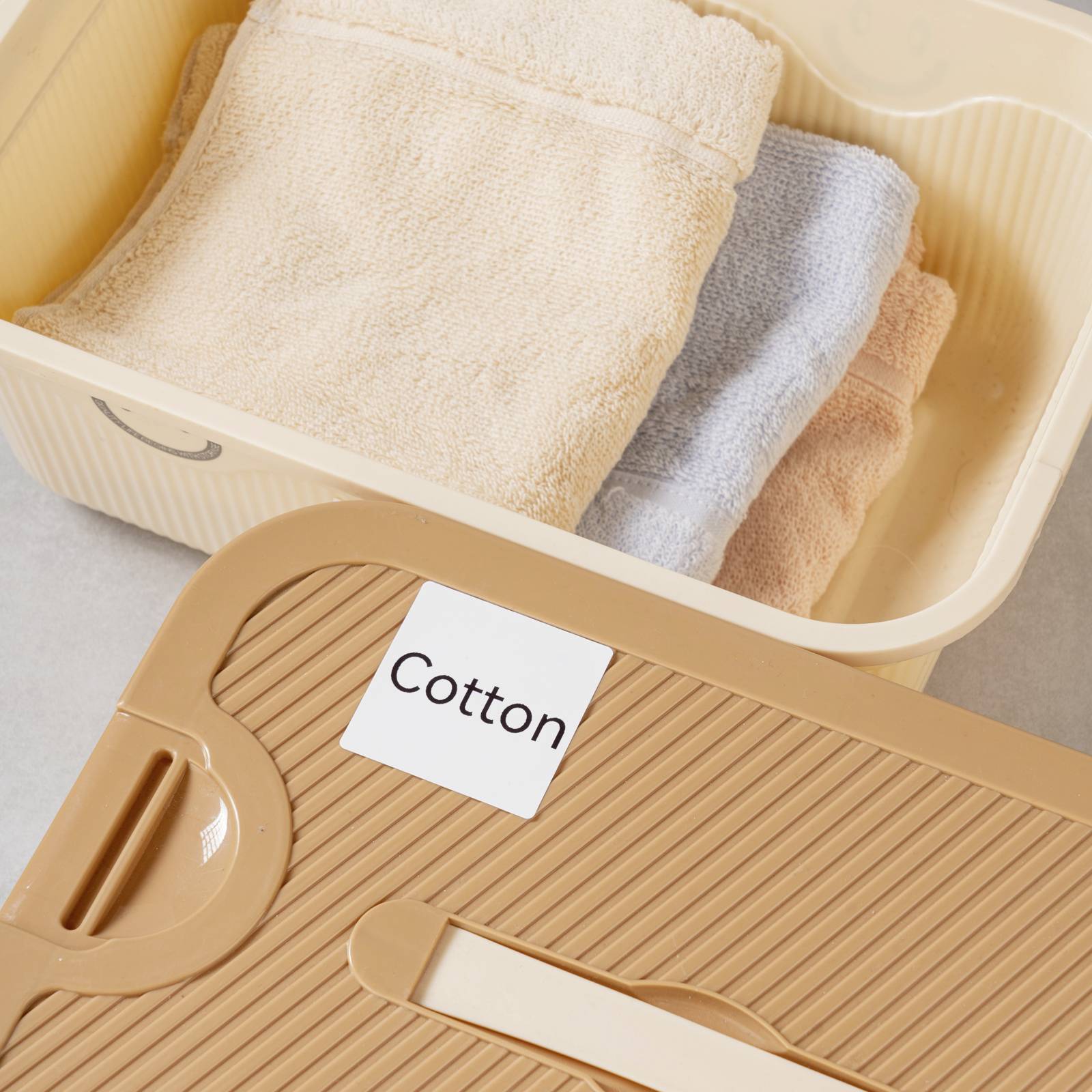 Kôš s poskladanými uterákmi, vrátane béžového, sivého a ďalšieho béžového. Vedľa leží veko s etiketou 'Cotton'.