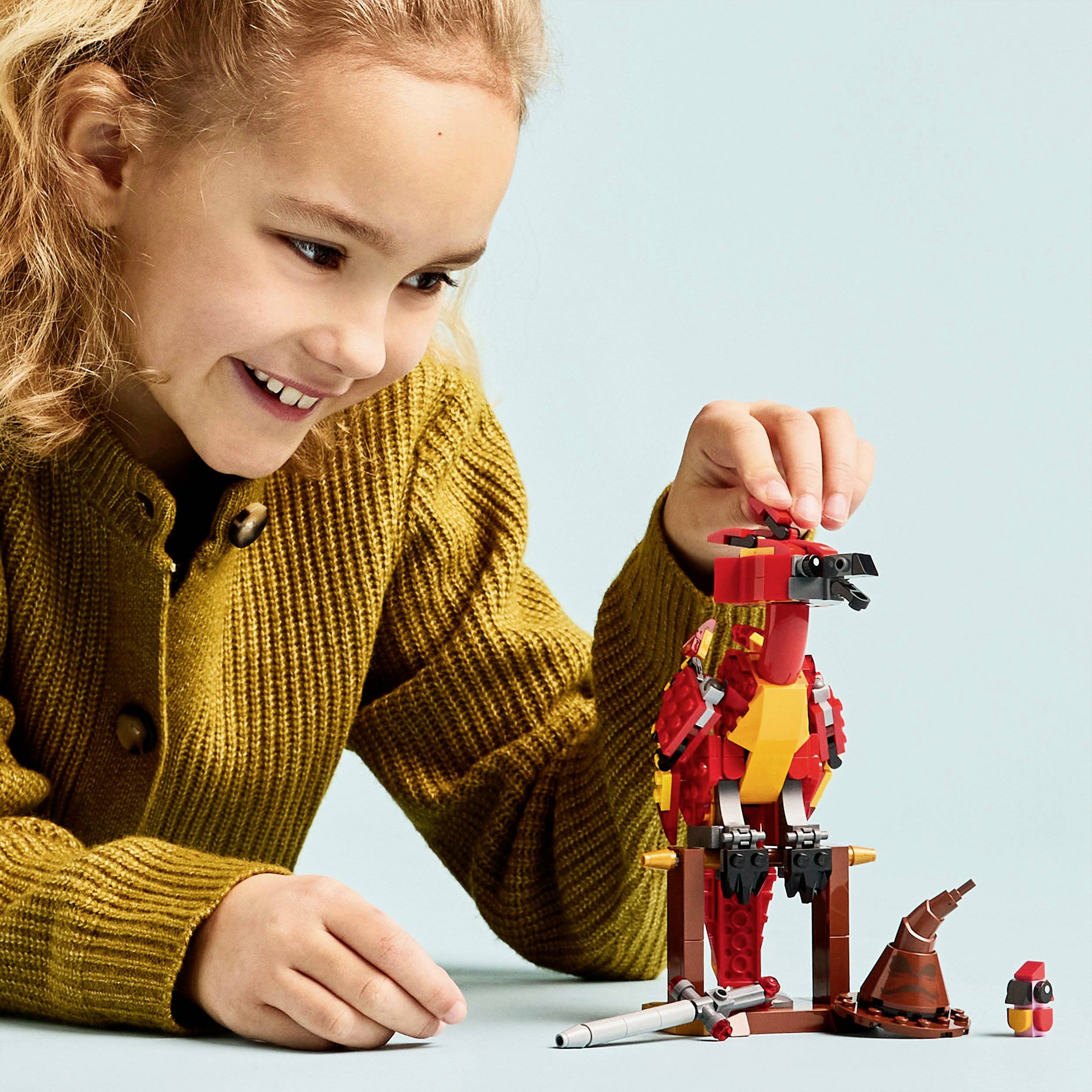 Usmievajúce sa dieťa v žltom svetri si hrá s červenou Lego figúrkou vtáka na modrom pozadí.