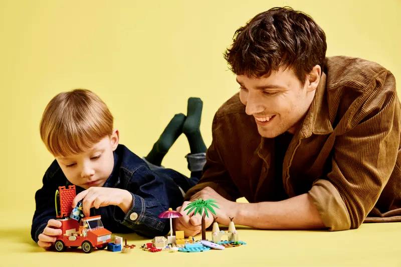 Dieťa sa hrá s hračkami LEGO s plážovou tematikou, pričom sa naň usmievajúci dospelý pozerá, v pozadí s žltým pozadím.