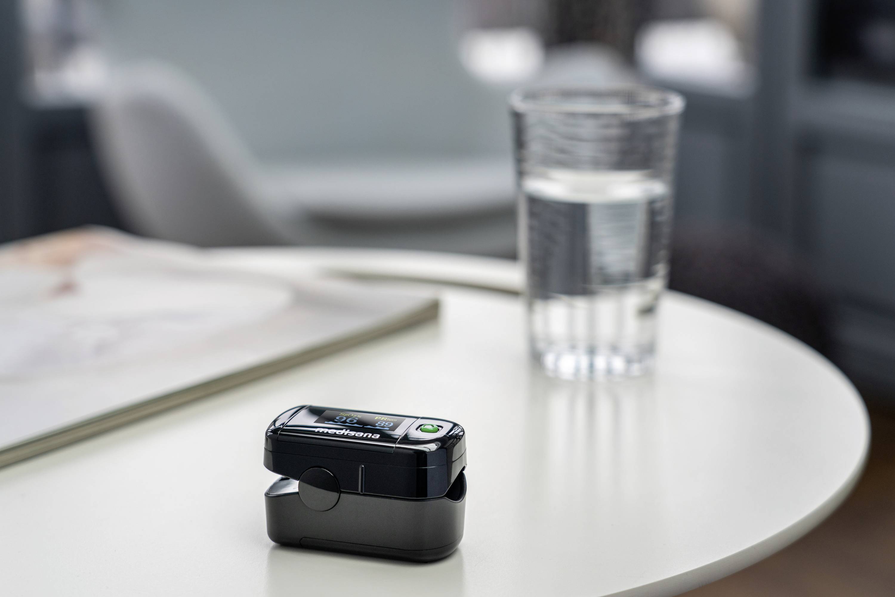 Pulzný oximeter leží na bielom stole vedľa pohára vody a kopy časopisov v svetlom interiéri.