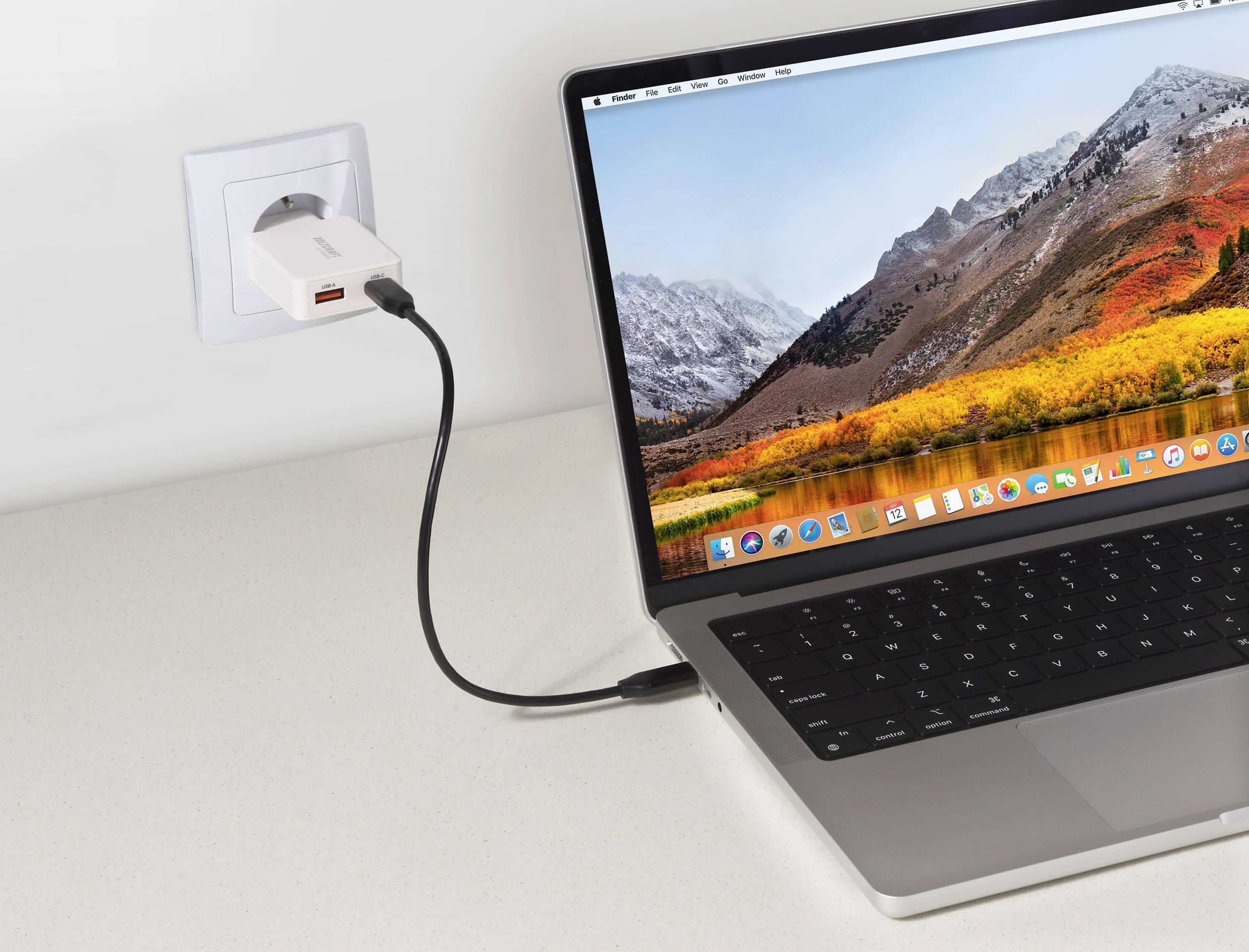 Laptop na stole sa nabíja, zapojený do elektrickej zásuvky na stene. Na obrazovke laptopa je tapeta s horskou krajinou a rôznymi ikonami aplikácií.