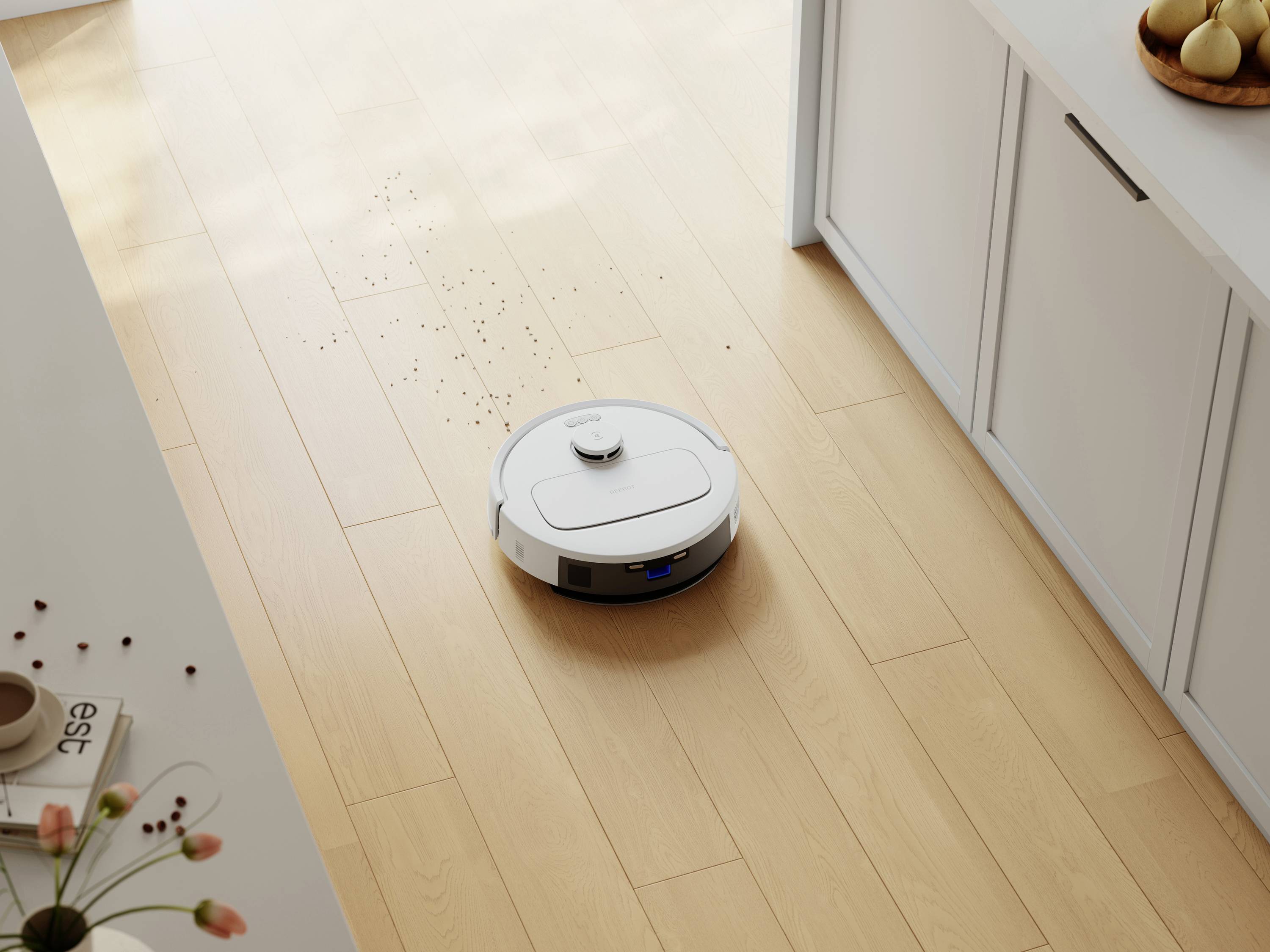 Biely robotický vysávač čistí drevené podlahu v kuchyni, na ktorej sú roztrúsené omrvinky. V pozadí sú viditeľné skrinky a miska s ovocím.