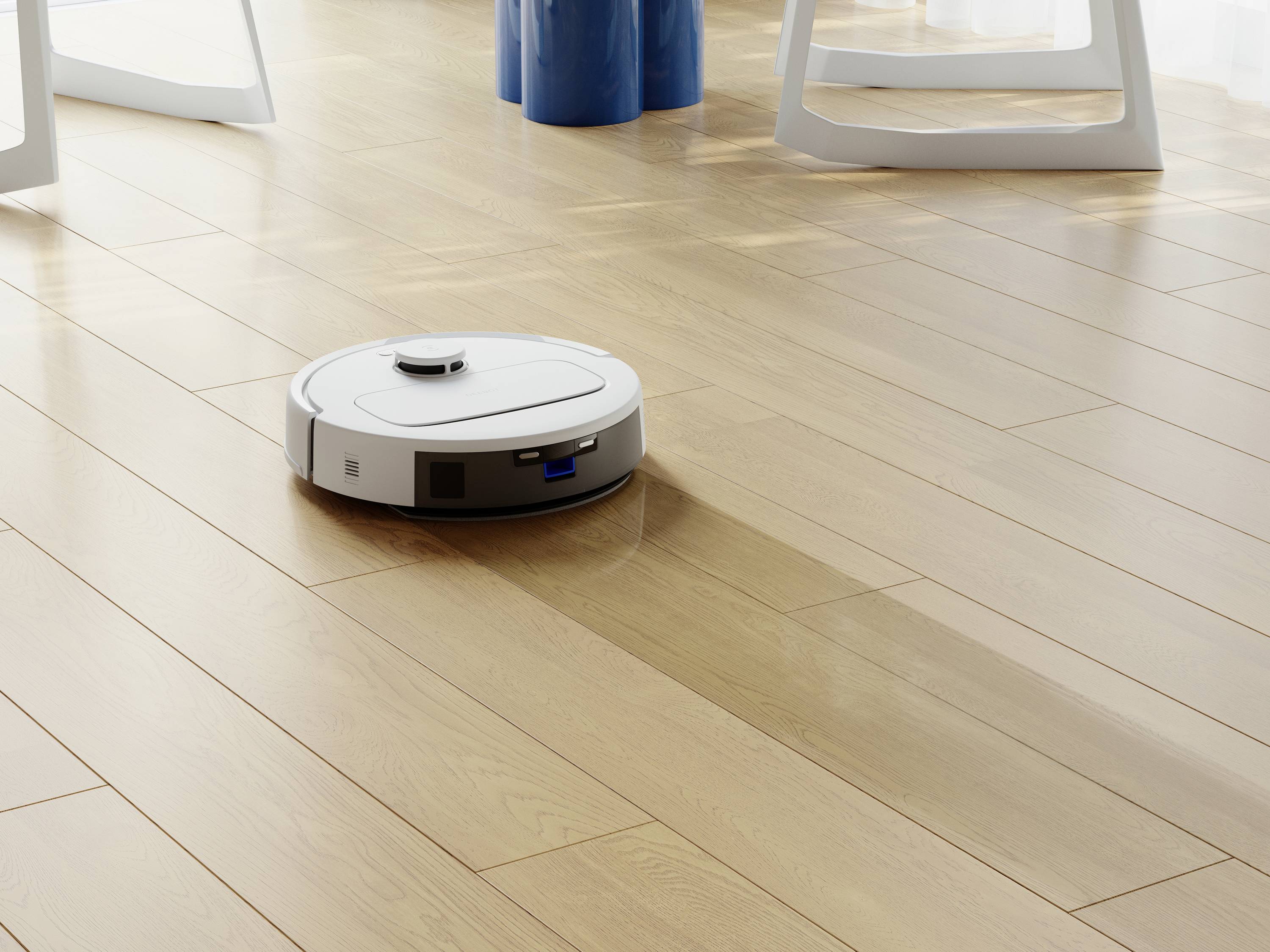 Biely robotický vysávač čistí drevenú podlahu v priestore zaplavenom slnečným svetlom. V pozadí sú viditeľné nohy stola.
