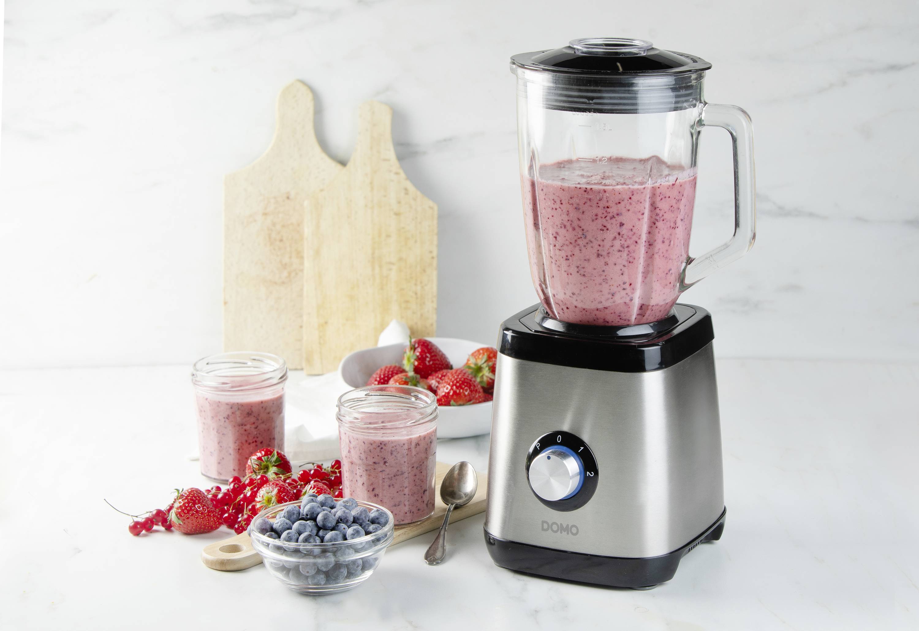 Stolový mixér obsahuje červený ovocný smoothie. Vedľa neho stoja naplnené poháre, čerstvé jahody, čučoriedky a lyžica.