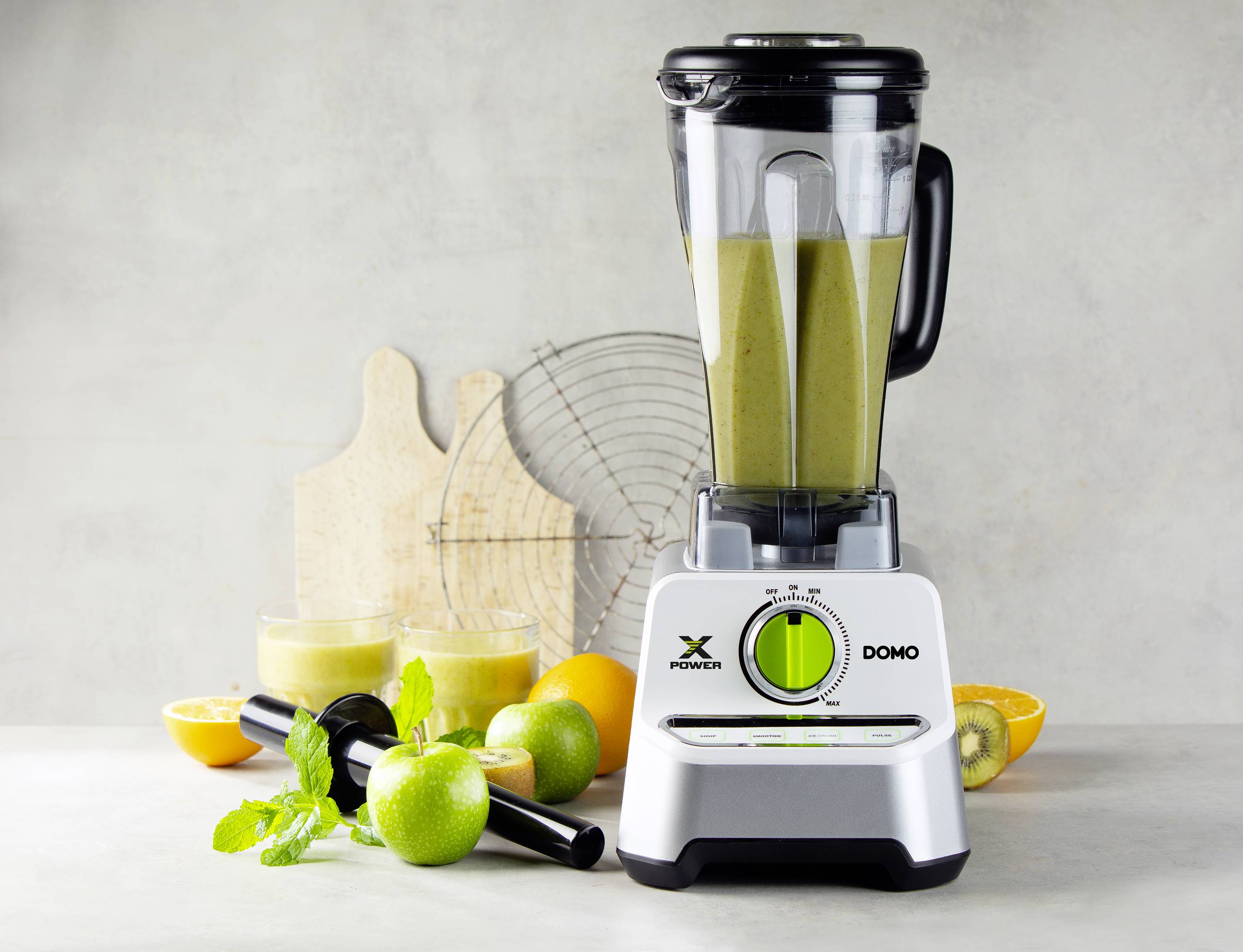 Mixér s naplnenou nádobou, obklopený limetkami, pomarančmi a kiwi. Dve sklá so smoothie a čerstvé listy mäty v popredí.