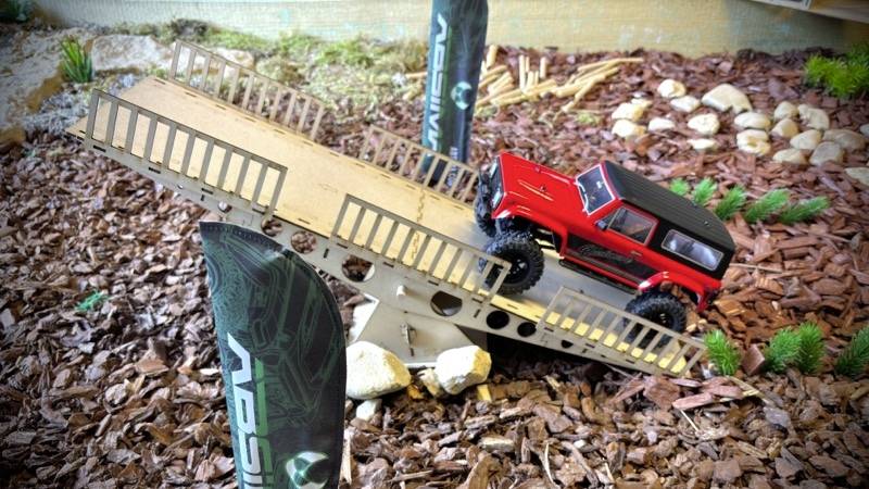 Červené modelové autíčko jazdí po šikmej drevenej rampe v miniatúrnej krajinke s drevennými štiepkami a malými rastlinami.