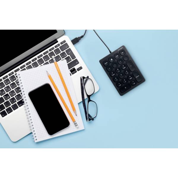 Laptop na modrom pozadí s poznámkovým blokom, smartfónom, ceruzkou, okuliarmi a externou numerickou klávesnicou vedľa.