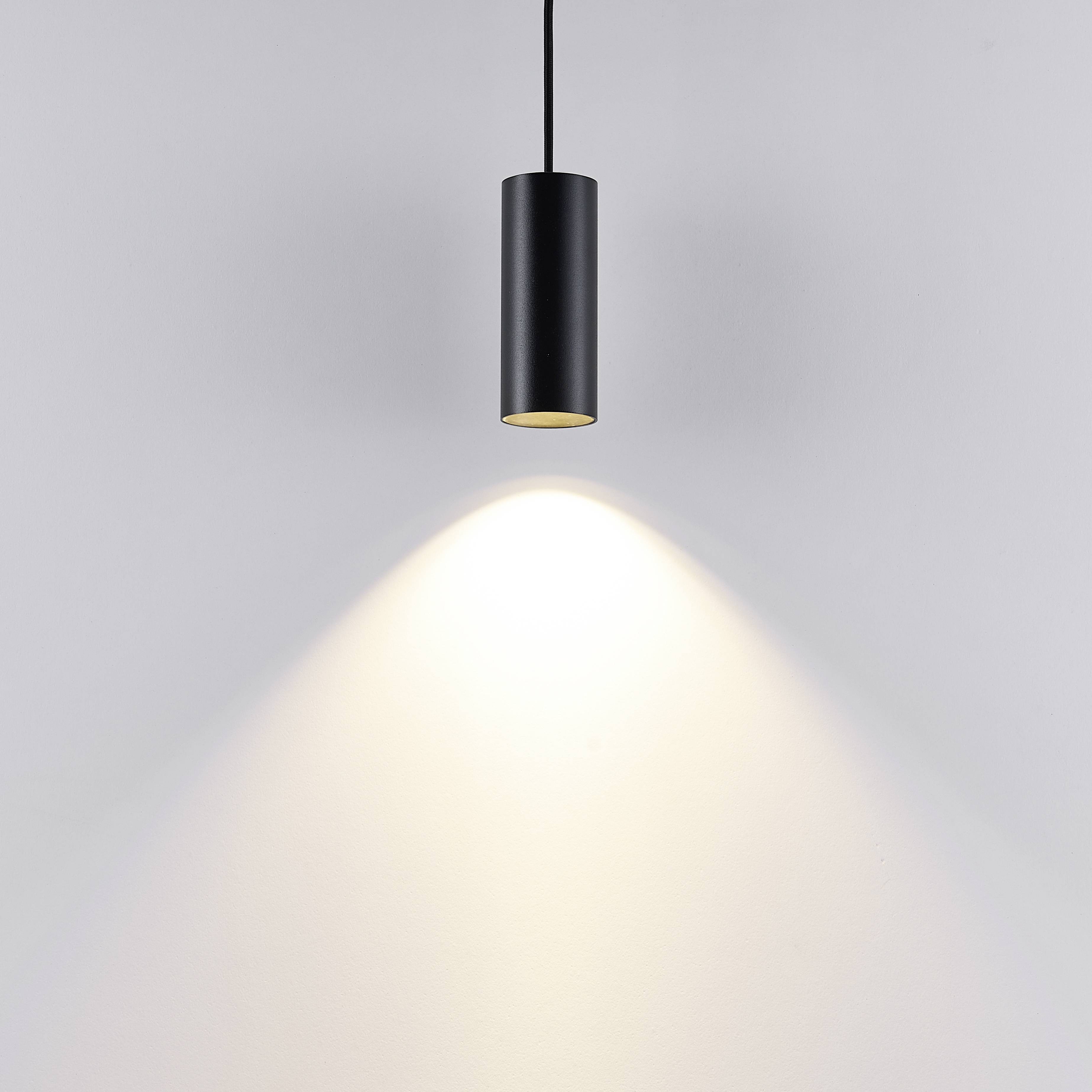 Minimalistická čierna závesná lampa, ktorá vrhá svetelný kužeľ na bielu stenu.