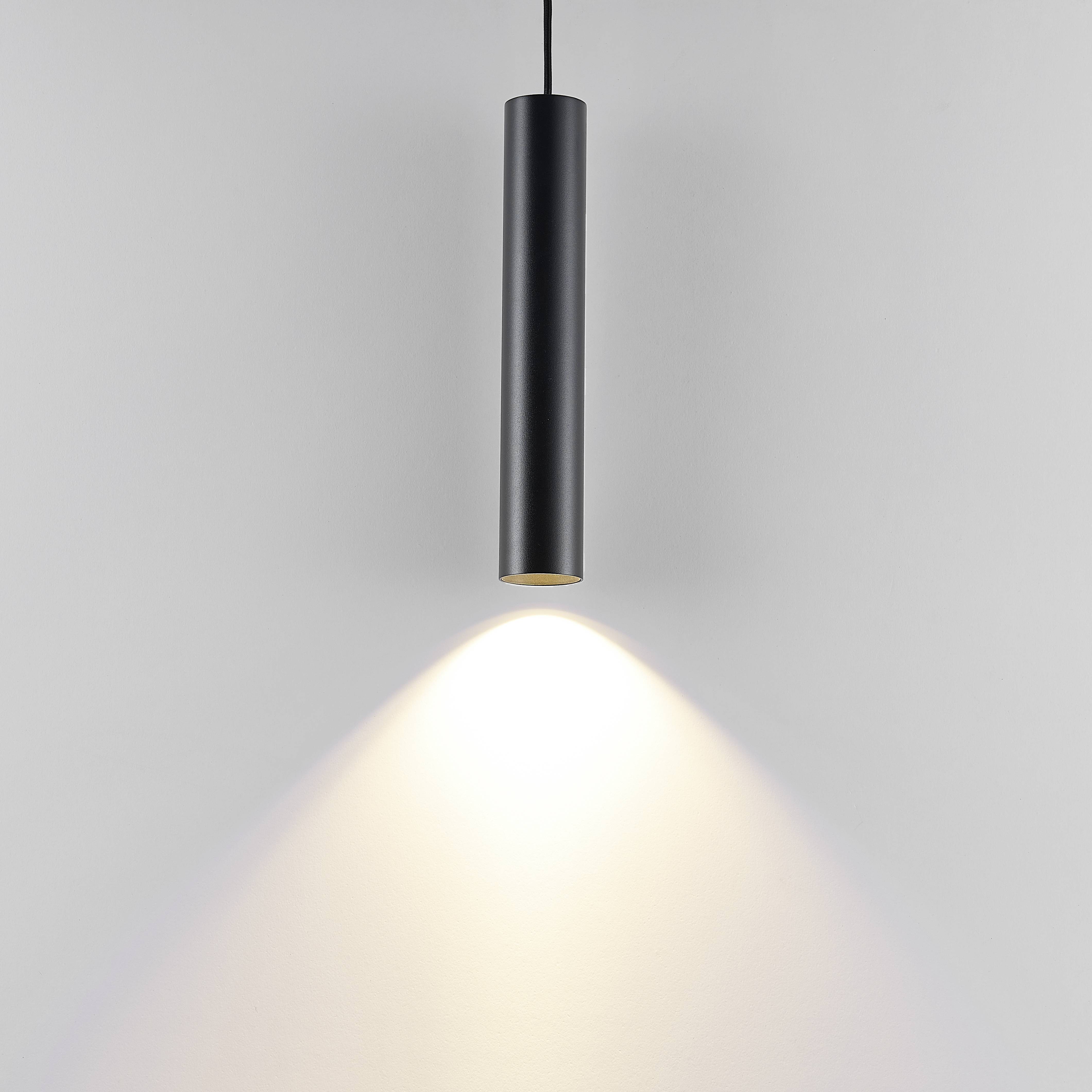 Závesná lampa s valcovým čiernym tienidlom, svieti smerom nadol a vytvára kužeľovitý svetelný lúč na sivej stene.