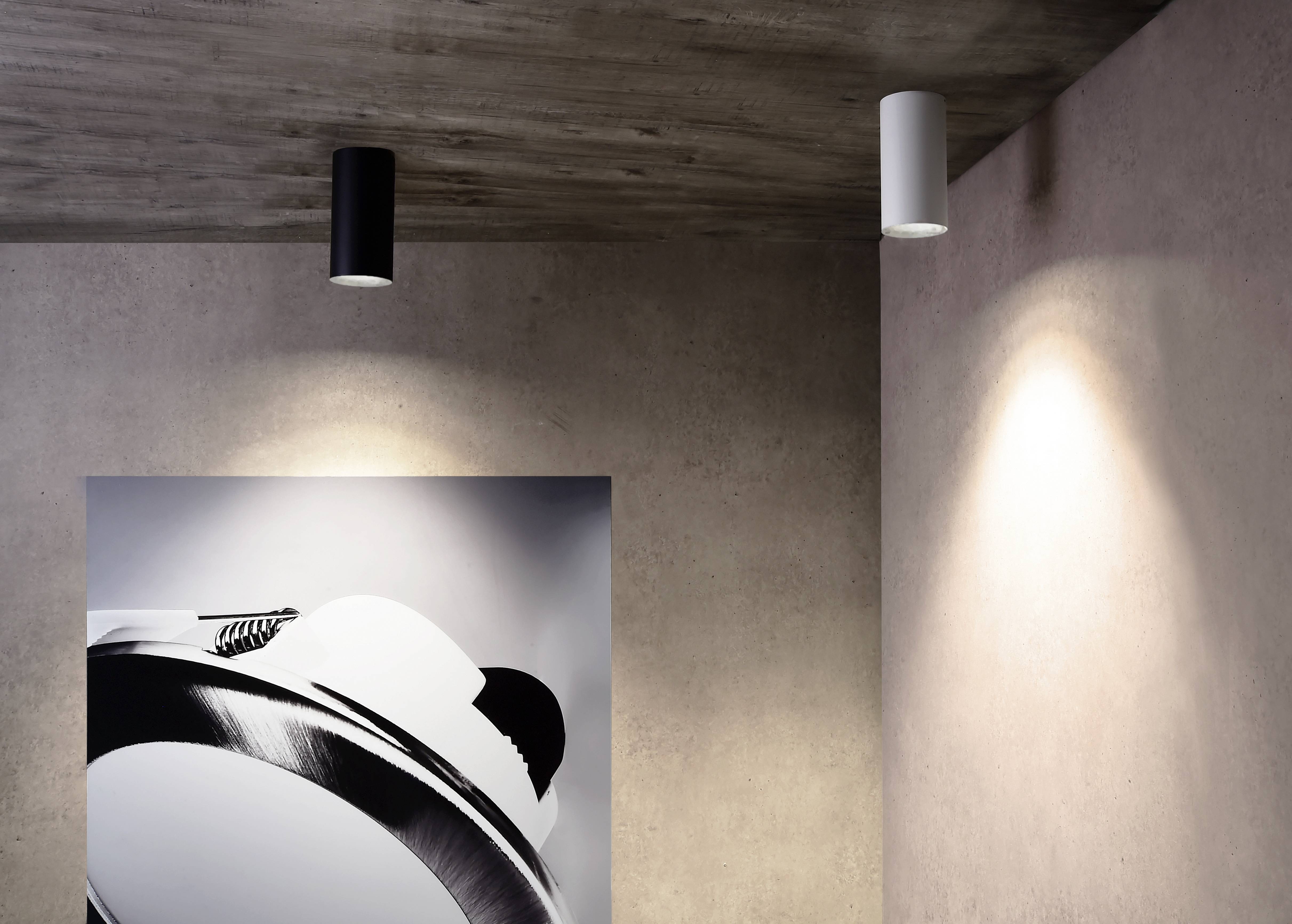 Dve valcové stropné svietidlá, jedno čierne a druhé biele, osvetľujú stenu s minimalistickým umeleckým dielom v čiernobielom prevedení.