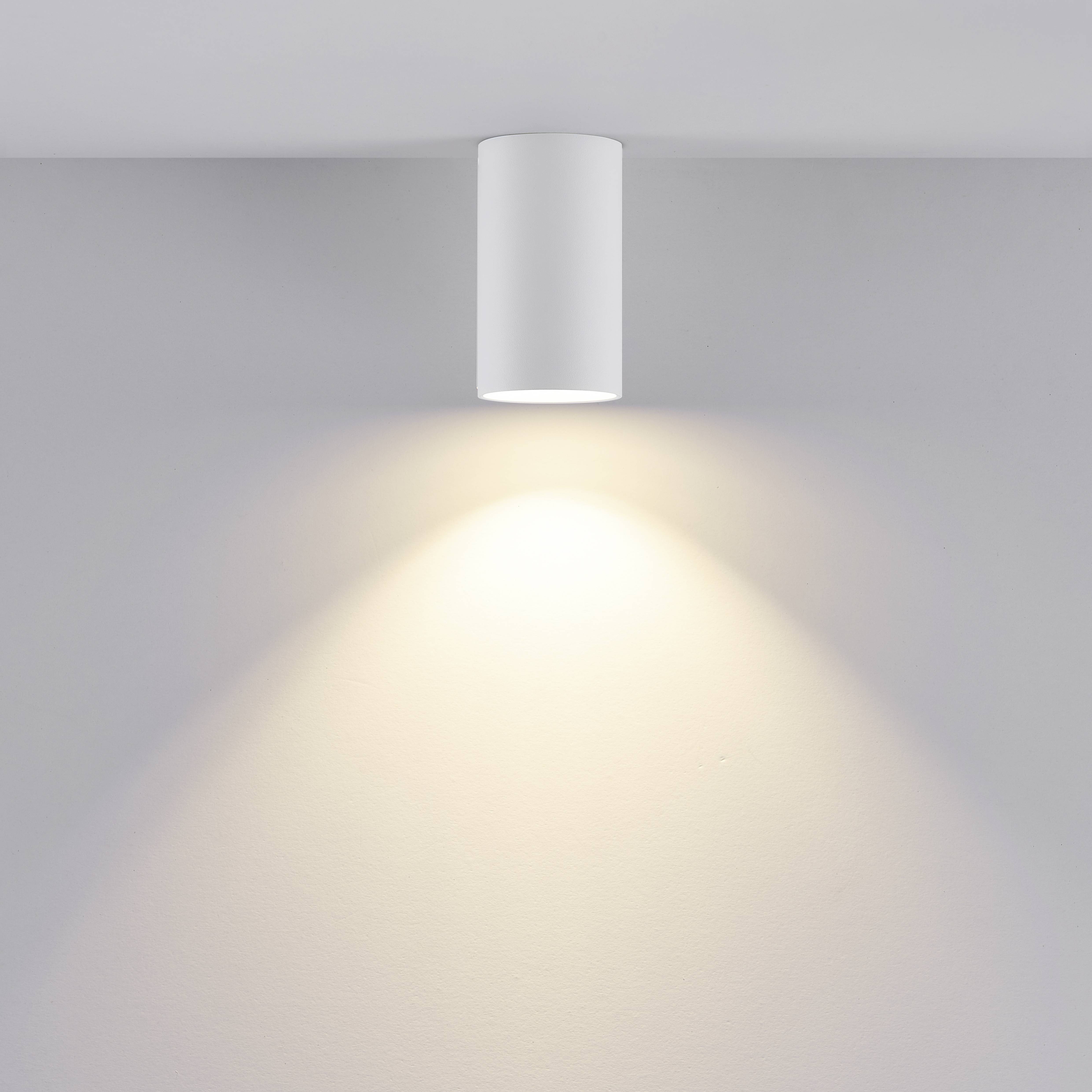 Biela valcovitá stropná lampa vyžaruje teplé svetlo na sivú stenu.