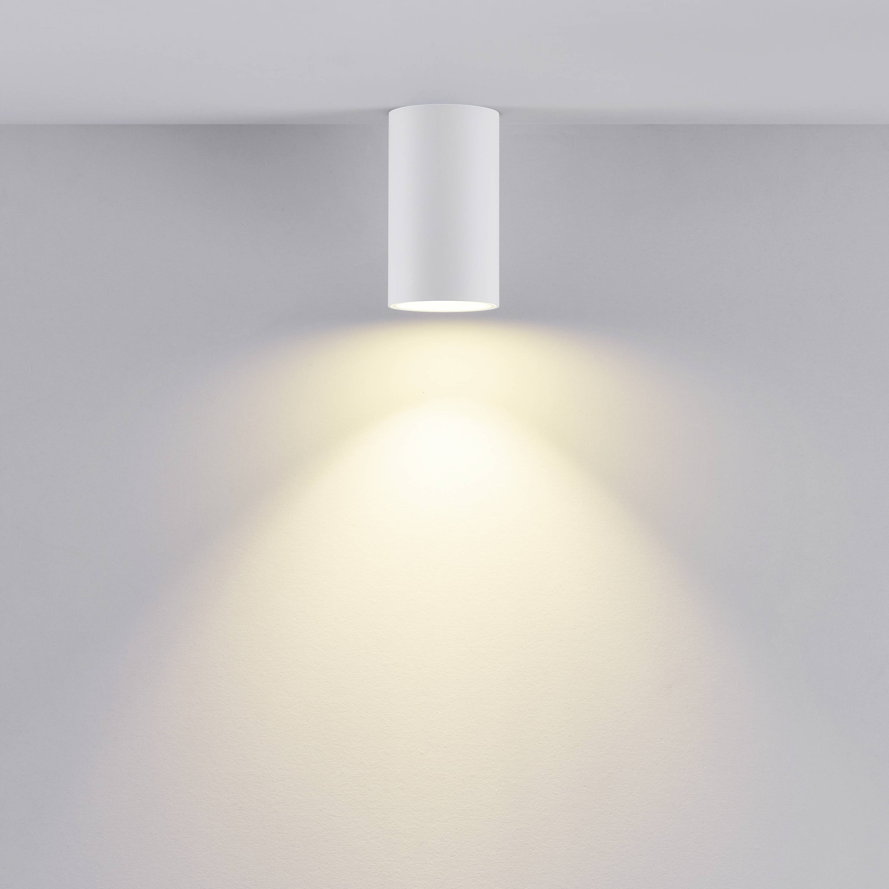 Valcové stropné svietidlo vyžaruje teplé svetlo na bielu stenu.