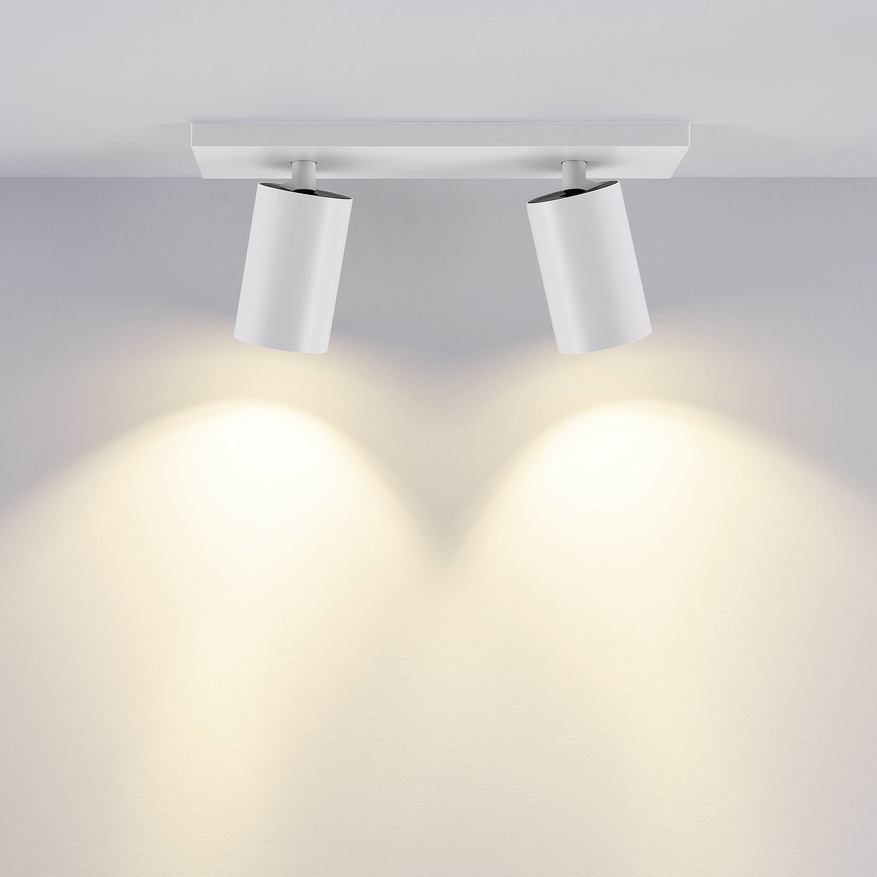 Dva valcové stropné svetlomety vrhajú teplé svetlo na bielu stenu.