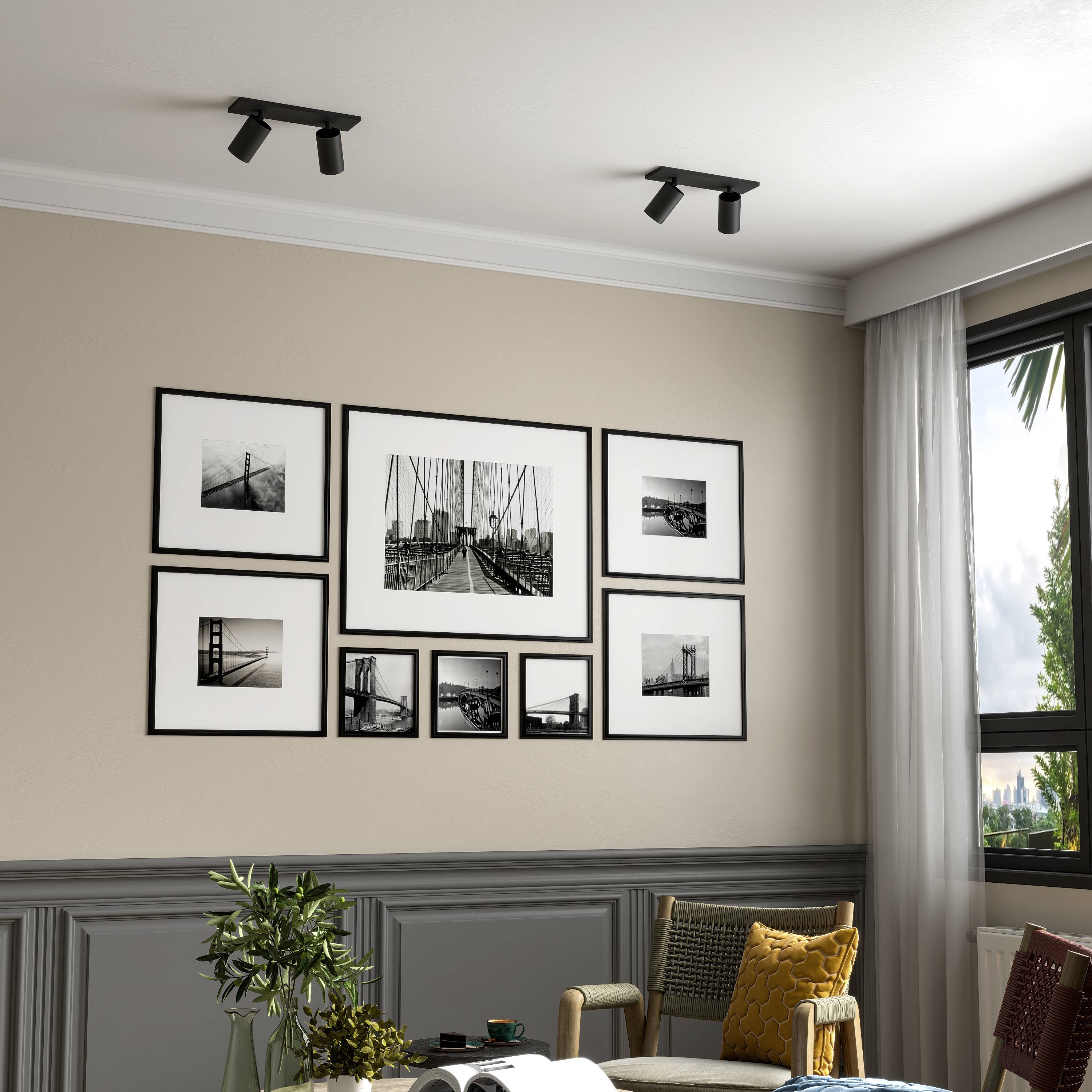 Obývacia izba s béžovou stenou a šedým obložením. Na stene visia rámované čiernobiele fotografie. Z okna sú vidieť zelené stromy vonku.