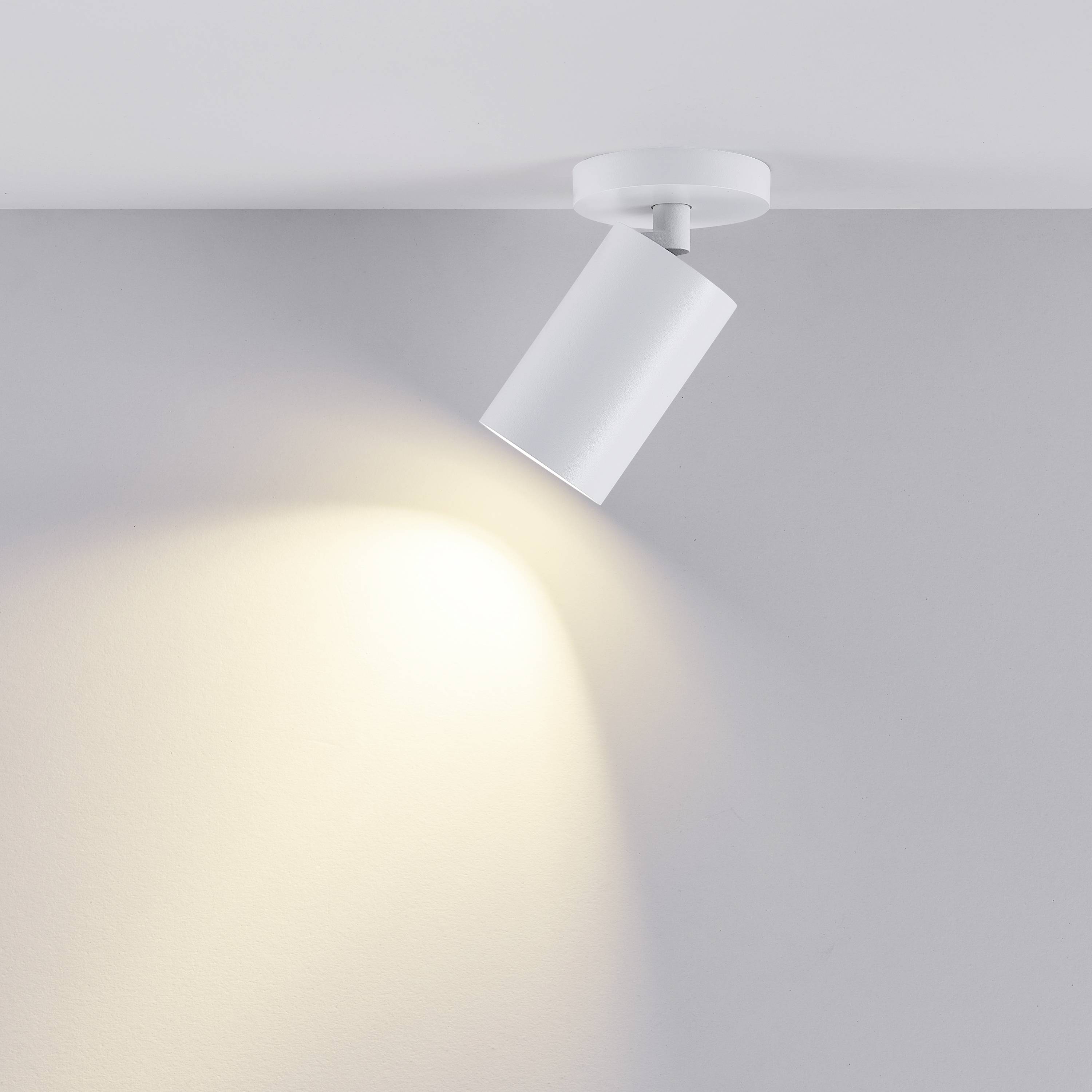 Biela stropná lampa s valcovitým dizajnom vyžaruje svetlo na svetlú stenu a vytvára teplú atmosféru.