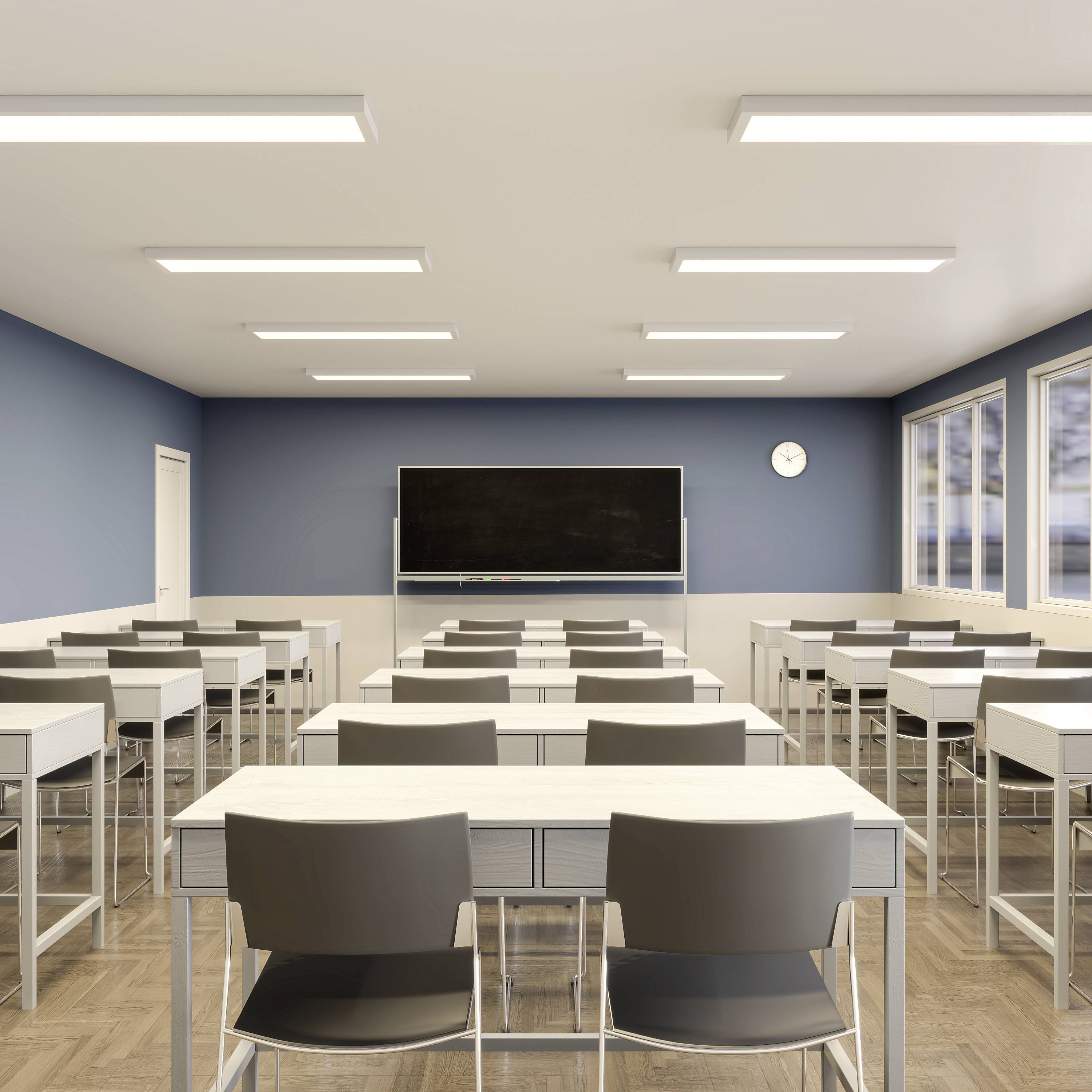 Prázdna učebňa so svetlými stenami a drevenou podlahou. Vpredu zelená tabuľa a nástenné hodiny. Rady stolov a stoličiek.